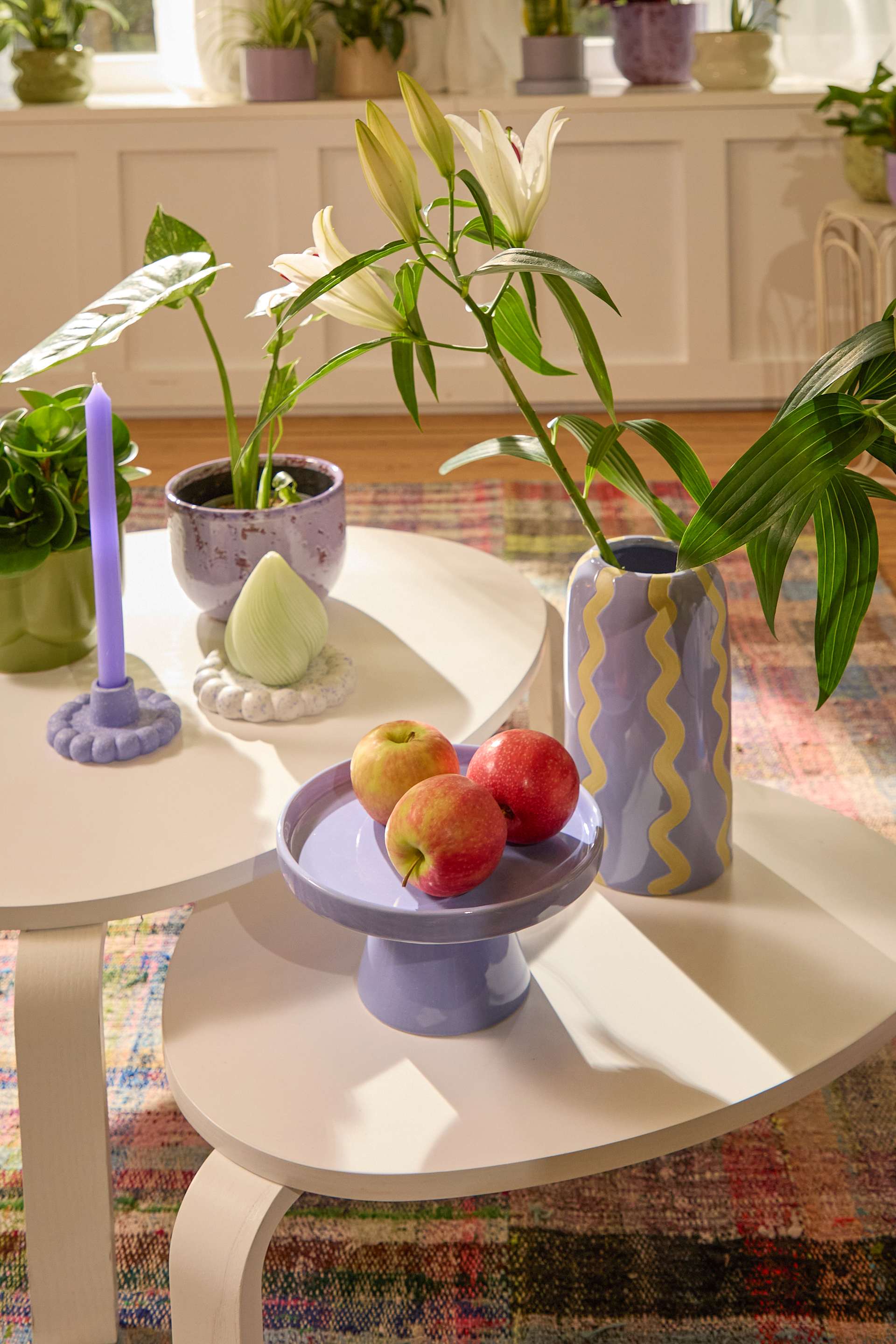 Eine gemütliche Atmosphäre in Innenräumen mit einem Tisch, auf dem verschiedene Dekorationselemente ausgestellt sind, darunter Topfpflanzen, eine Kerze und ein Teller mit frischem Obst.