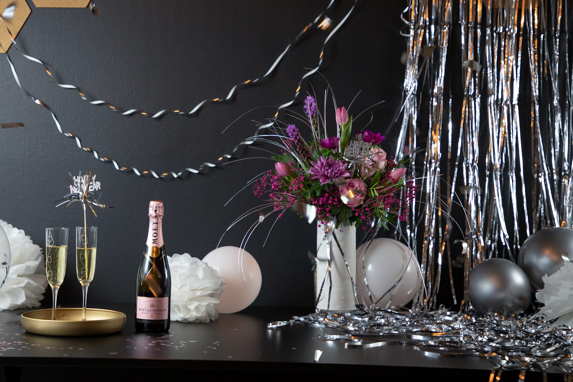 Eine festlich gedeckte Tafel mit einem Strauß leuchtender Blumen, Champagnergläsern und einer Vielzahl dekorativer Elemente, alles vor einem dunklen Hintergrund mit schimmernden metallischen Akzenten.