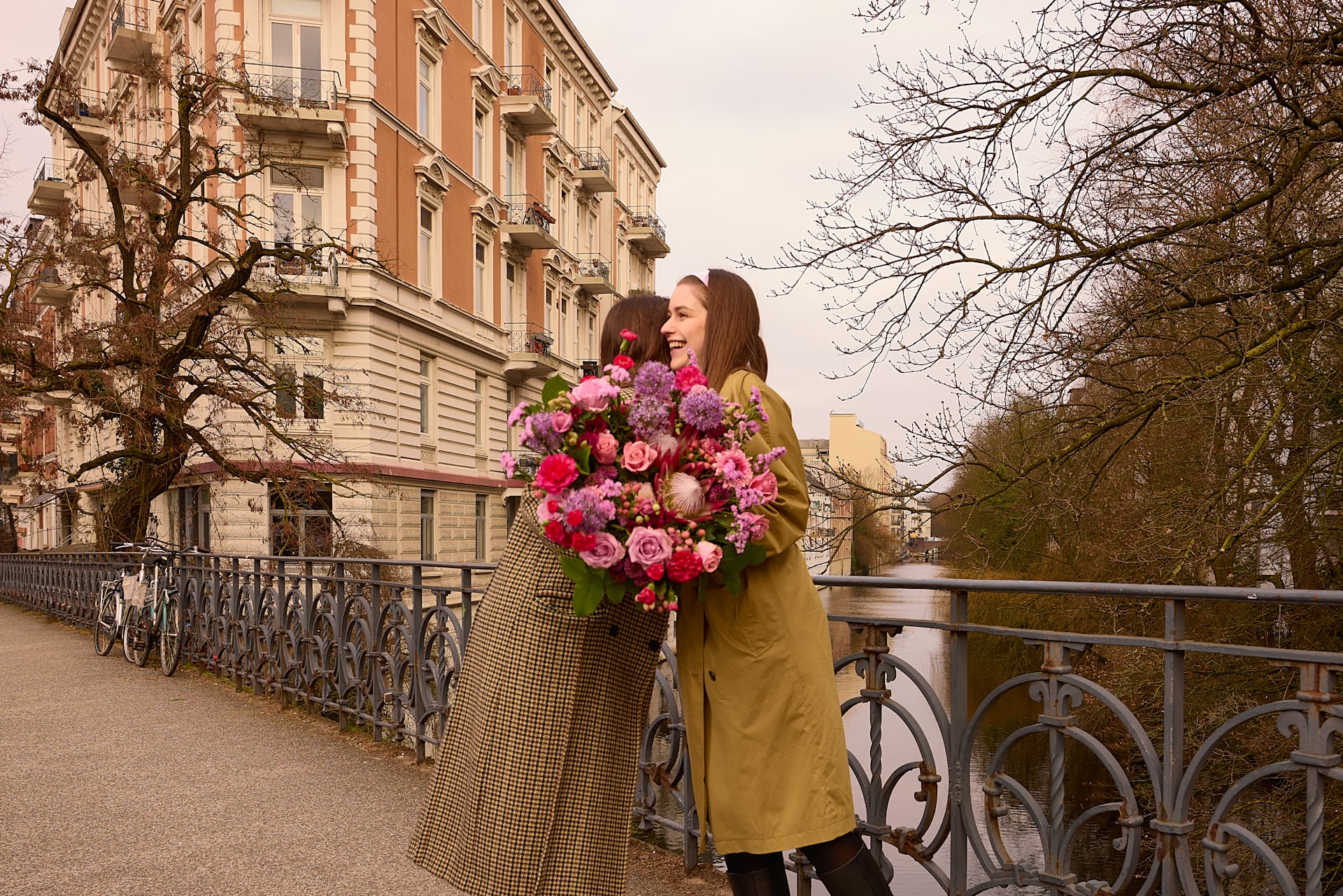 Zwei junge Frauen in farbenfrohen Mänteln stehen auf einer Brücke und halten einen großen Strauß leuchtend pinkfarbener Blumen. Im Hintergrund sind ein historisches Gebäude und kahle Bäume zu sehen.