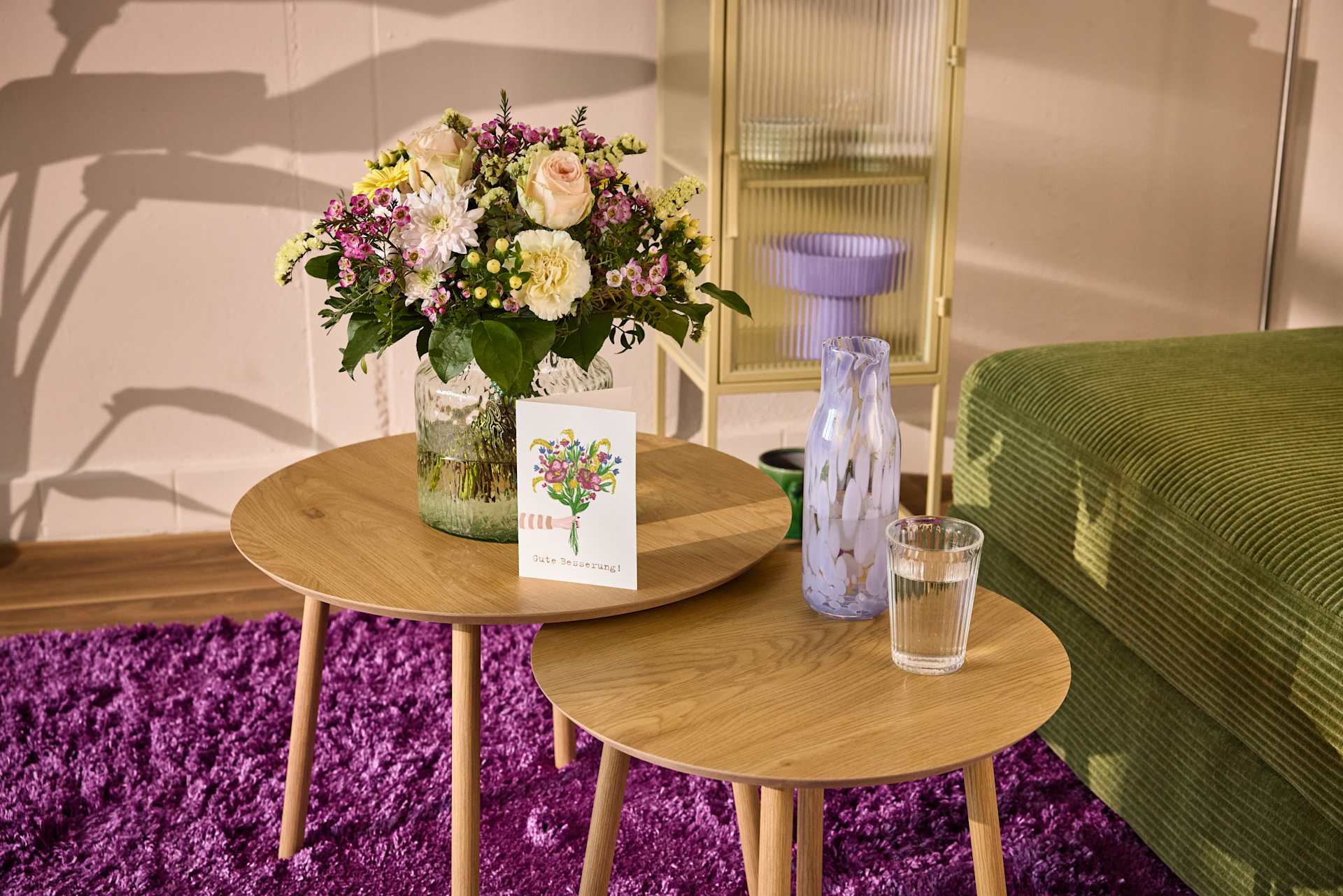 Auf einem Holztisch steht ein farbenprächtiger Blumenstrauß, umgeben von einem flauschigen lila Teppich und einem grünen Sofa im Hintergrund.