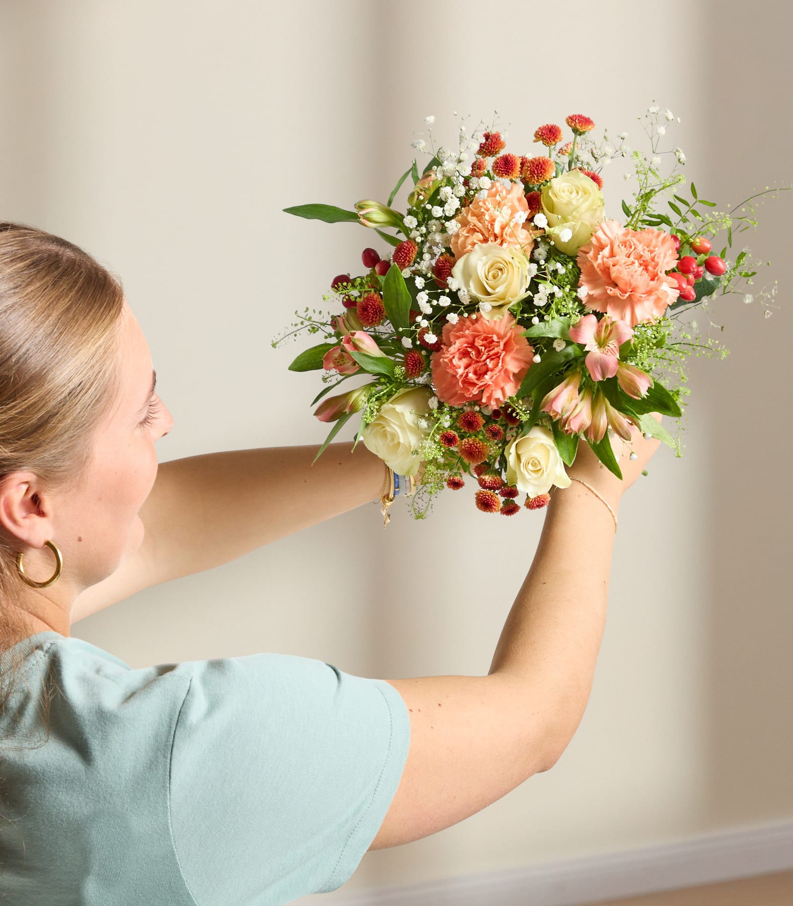 Ein leuchtender Strauß gelber Rosen, rosa Nelken und anderer bunter Blumen in einer grünen Glasvase steht auf einer hellen Fläche vor einem schlichten Hintergrund.