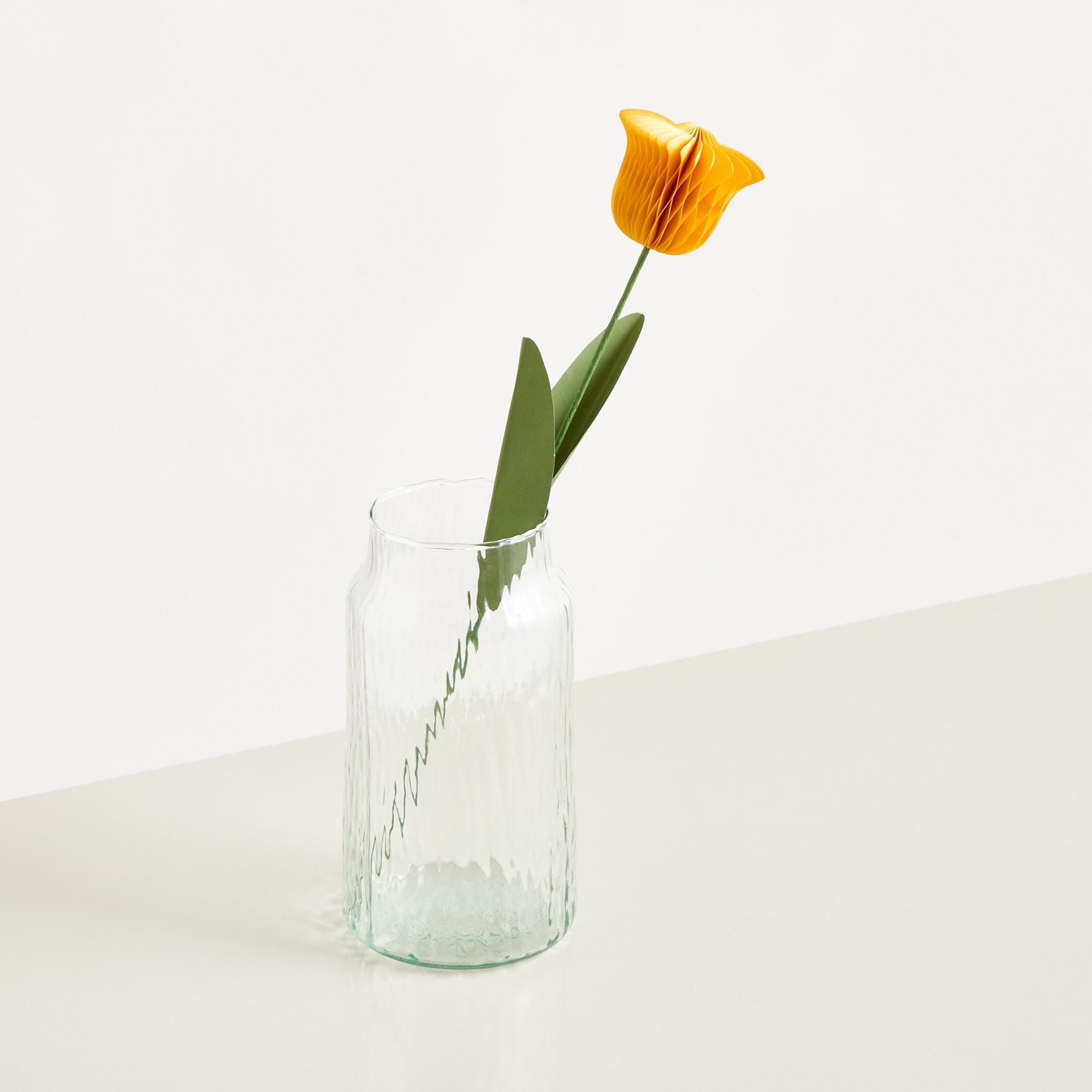 Eine einzelne, leuchtend orangefarbene Tulpe steht in einer durchsichtigen Glasvase vor einem schlichten weißen Hintergrund.