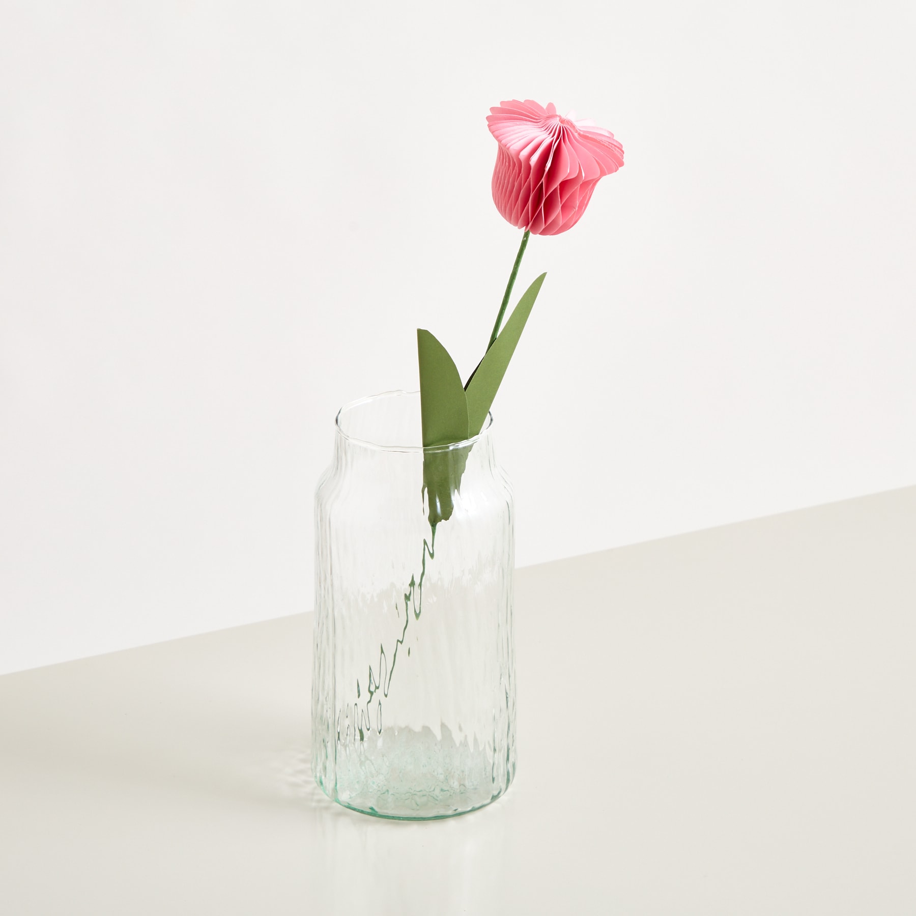 Eine einzelne rosa Tulpe steht in einer durchsichtigen Glasvase vor einem schlichten weißen Hintergrund.