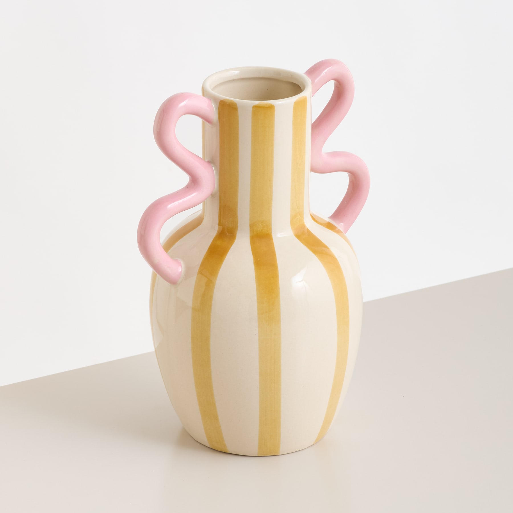 Eine gelb-weiß gestreifte Keramikvase mit rosa Henkeln steht auf einer neutralfarbenen Fläche vor einem schlichten Hintergrund.
