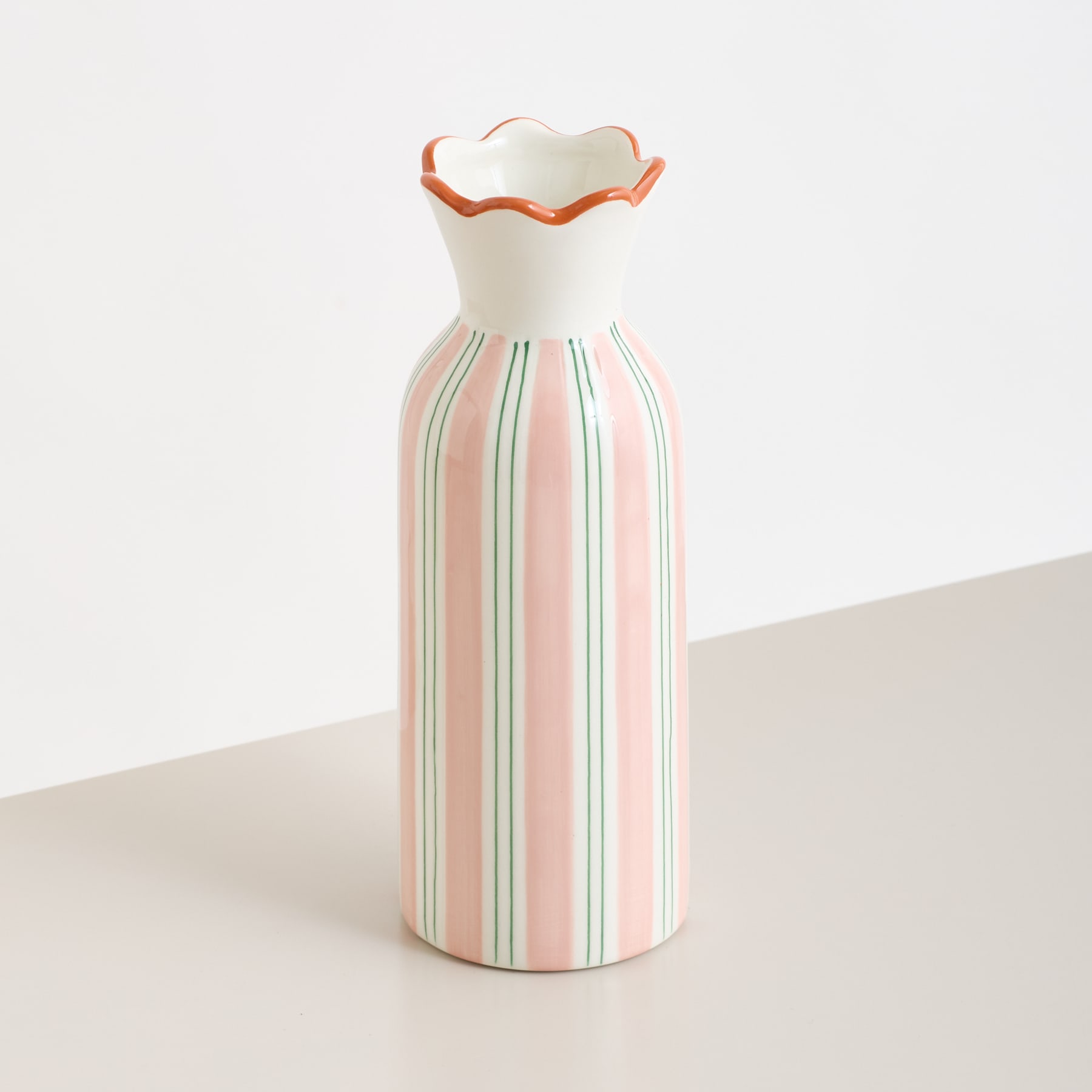 Eine weiße Keramikvase mit gewelltem Rand und vertikalen rosa, grünen und orangen Streifen steht auf einer hellen Fläche vor einem schlichten Hintergrund.