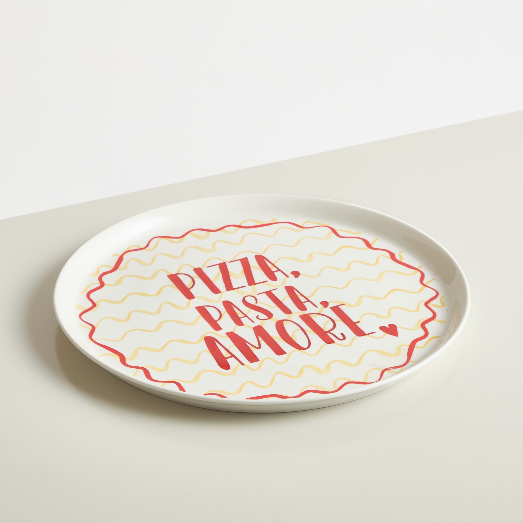 Ein weißer Keramikteller mit der Aufschrift „Pizza, pasta, amore“ in roter und gelber Schreibschrift vor einem schlichten weißen Hintergrund.