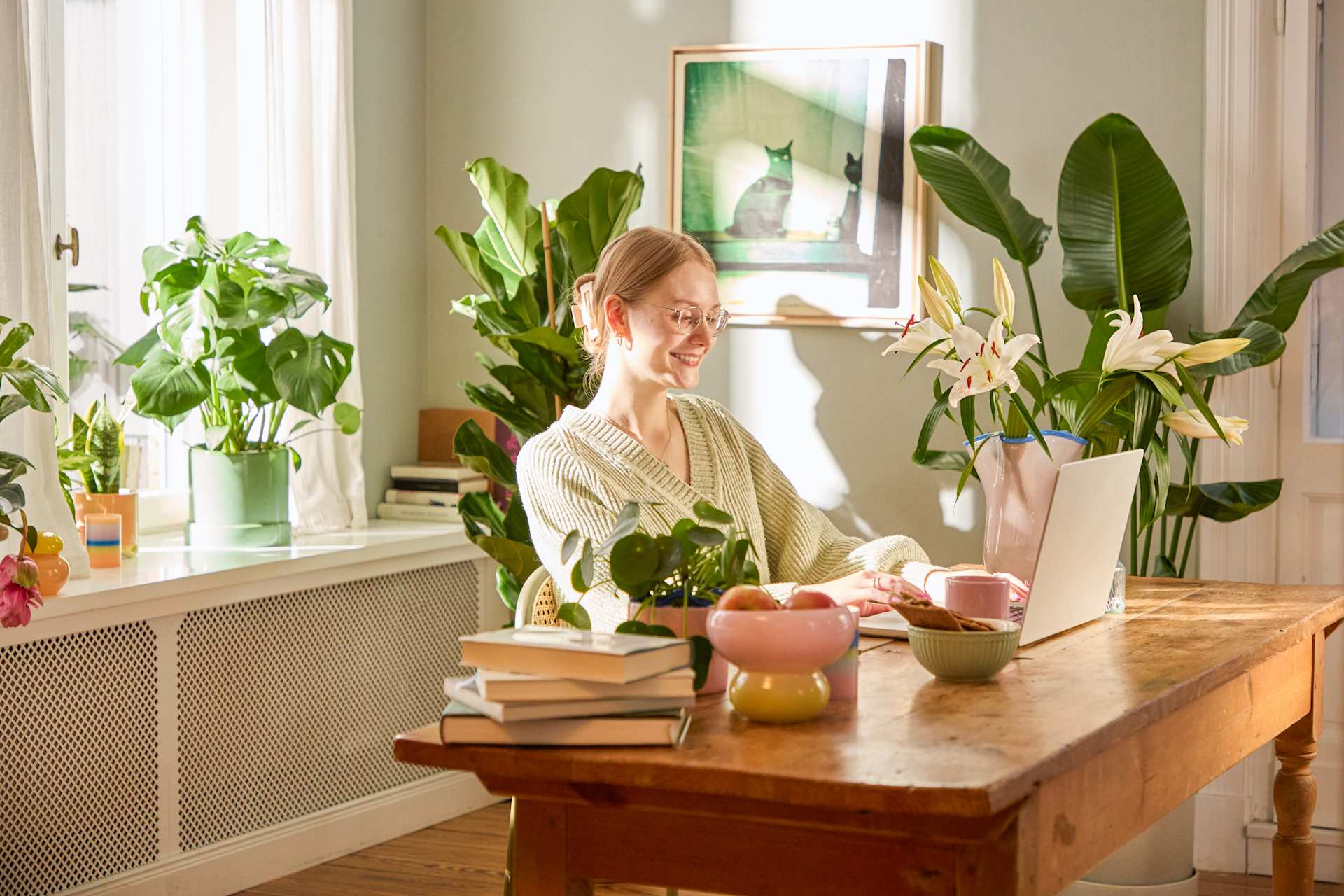 Ein gemütliches und einladendes Wohninterieur mit einer Frau, die an einem Holztisch sitzt, umgeben von üppigen Zimmerpflanzen und Büchern.
