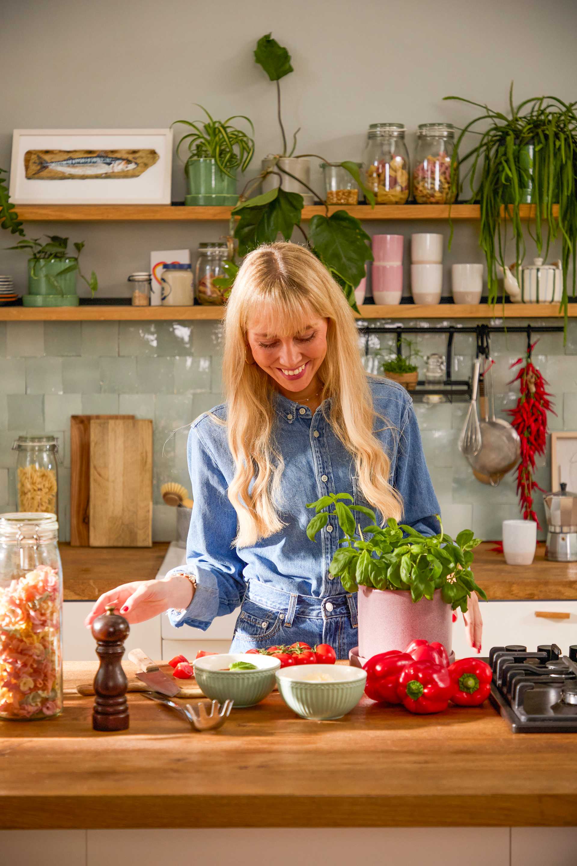 Eine lächelnde Frau mit langen blonden Haaren bereitet Essen an einer Küchentheke zu, die von verschiedenen Kochutensilien, Pflanzen und Regalen voller Gläser und Geschirr umgeben ist.