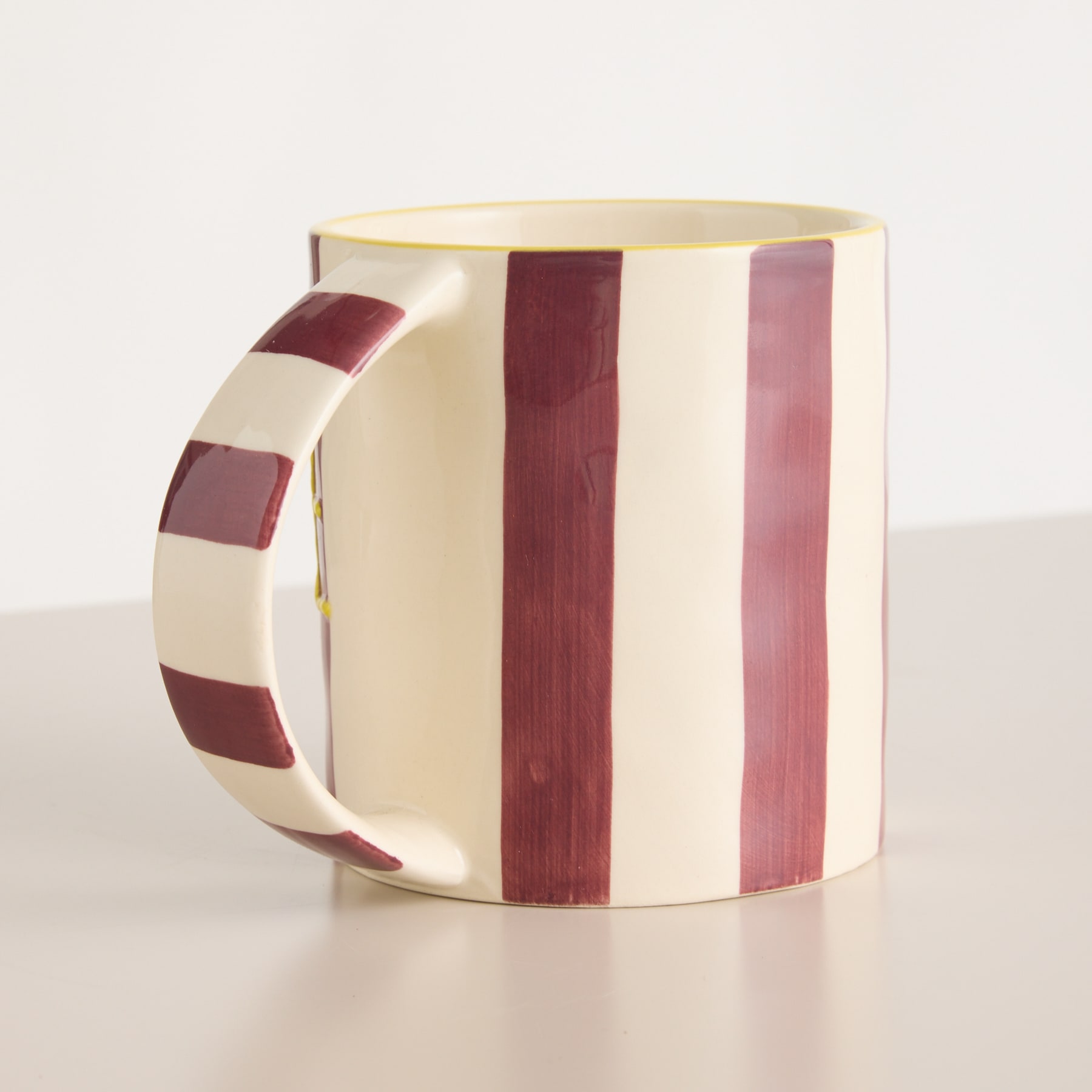 Ein Keramikbecher mit Streifenmuster in Creme-, Burgunder- und Rosatönen steht auf einer hellen Oberfläche.