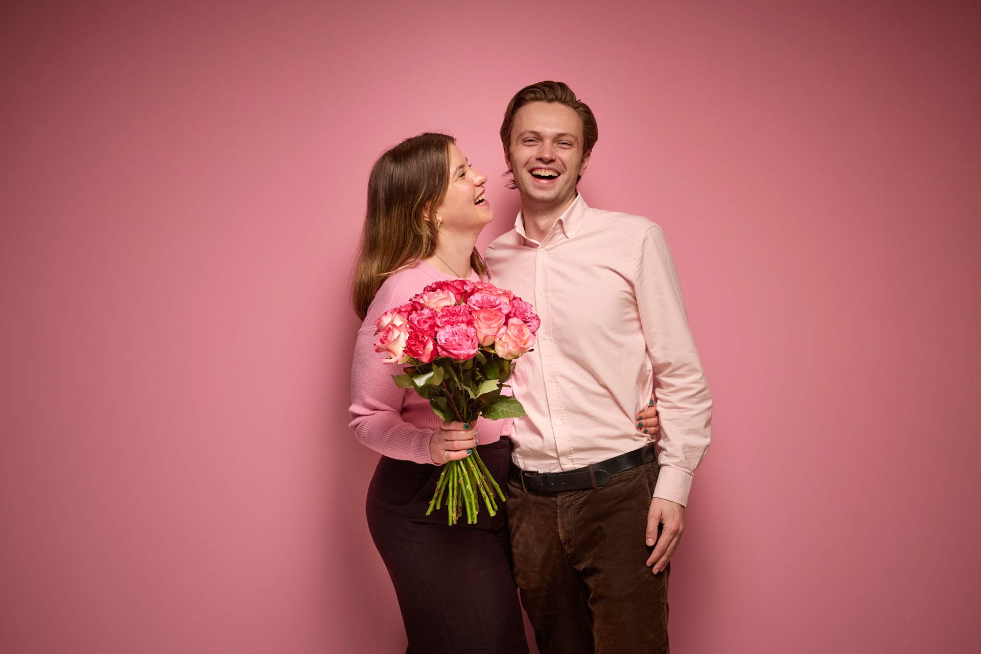 Ein junges Paar steht zusammen, die Frau hält einen Strauß rosa Rosen vor einem leuchtend rosa Hintergrund.