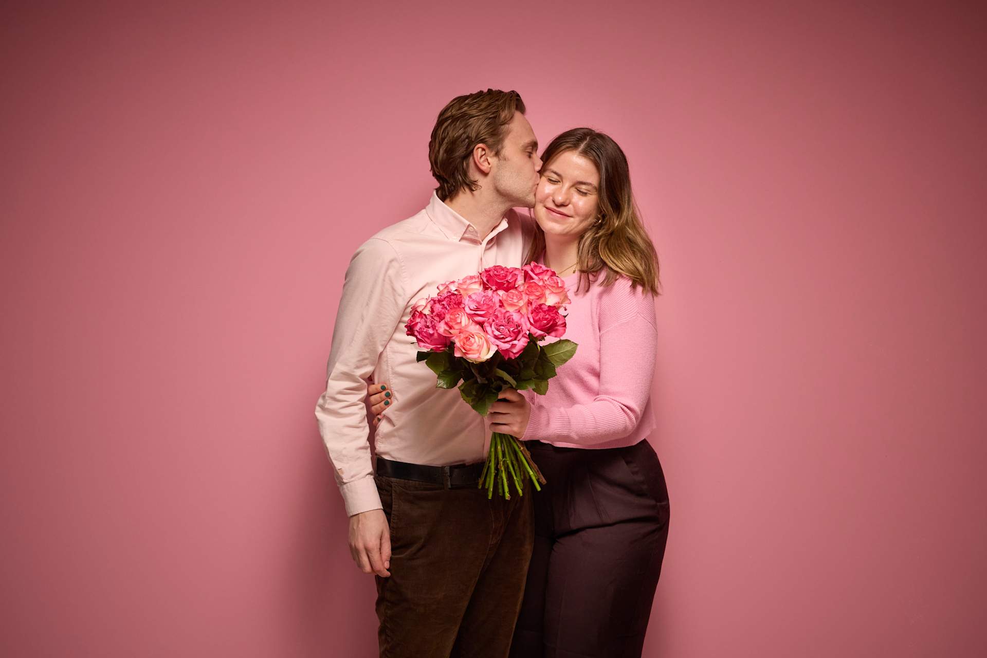 Ein Paar umarmt sich und hält einen Strauß rosa Nelken vor einem einfarbig rosa Hintergrund.