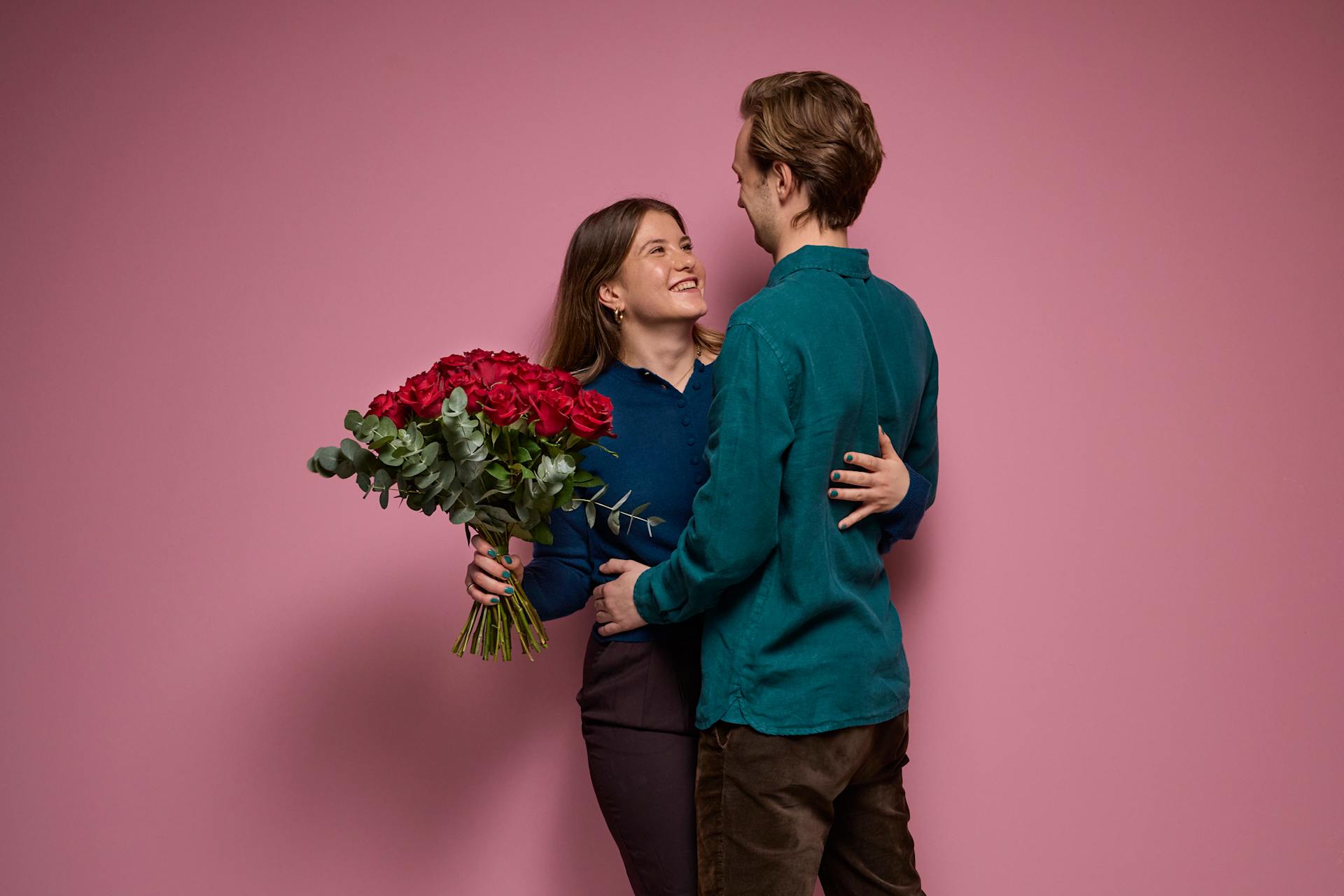 Ein junges Paar umarmt sich, die Frau hält einen Strauß roter Rosen vor einem leuchtend pinkfarbenen Hintergrund.