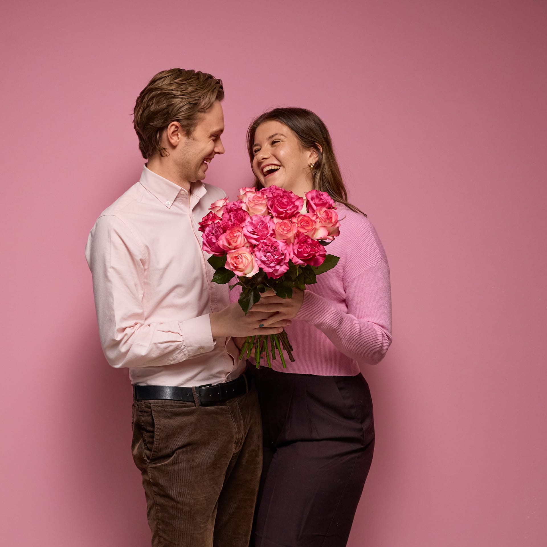 Ein junges Paar steht zusammen, die Frau hält einen Strauß leuchtend pinkfarbener Rosen, beide lächeln freudig vor einem einfarbig pinkfarbenen Hintergrund.