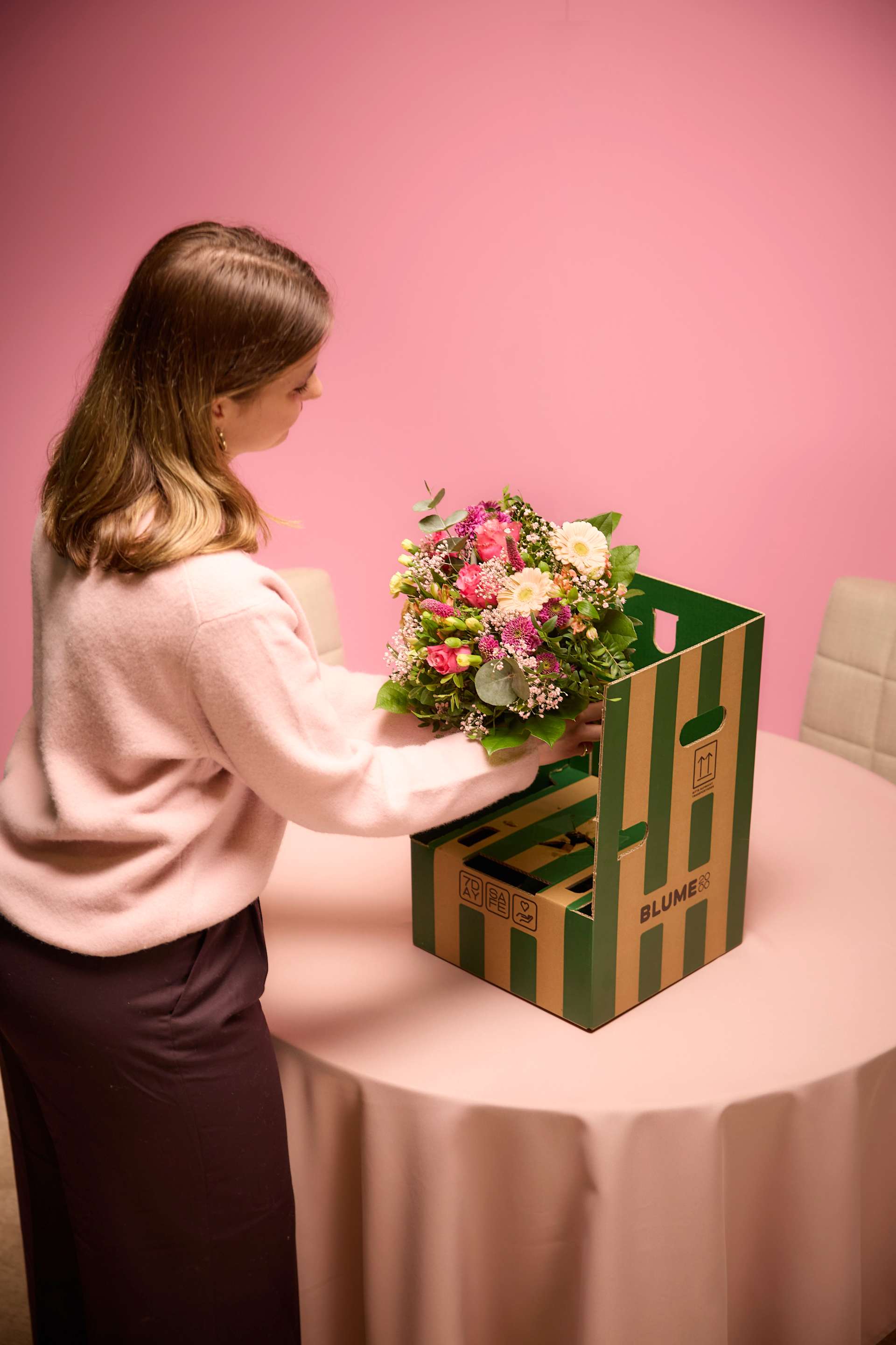 Eine Frau in einem rosa Pullover hält einen Blumenstrauß vor einer grün-weiß gestreiften Schachtel auf einem Tisch vor einem rosa Hintergrund.