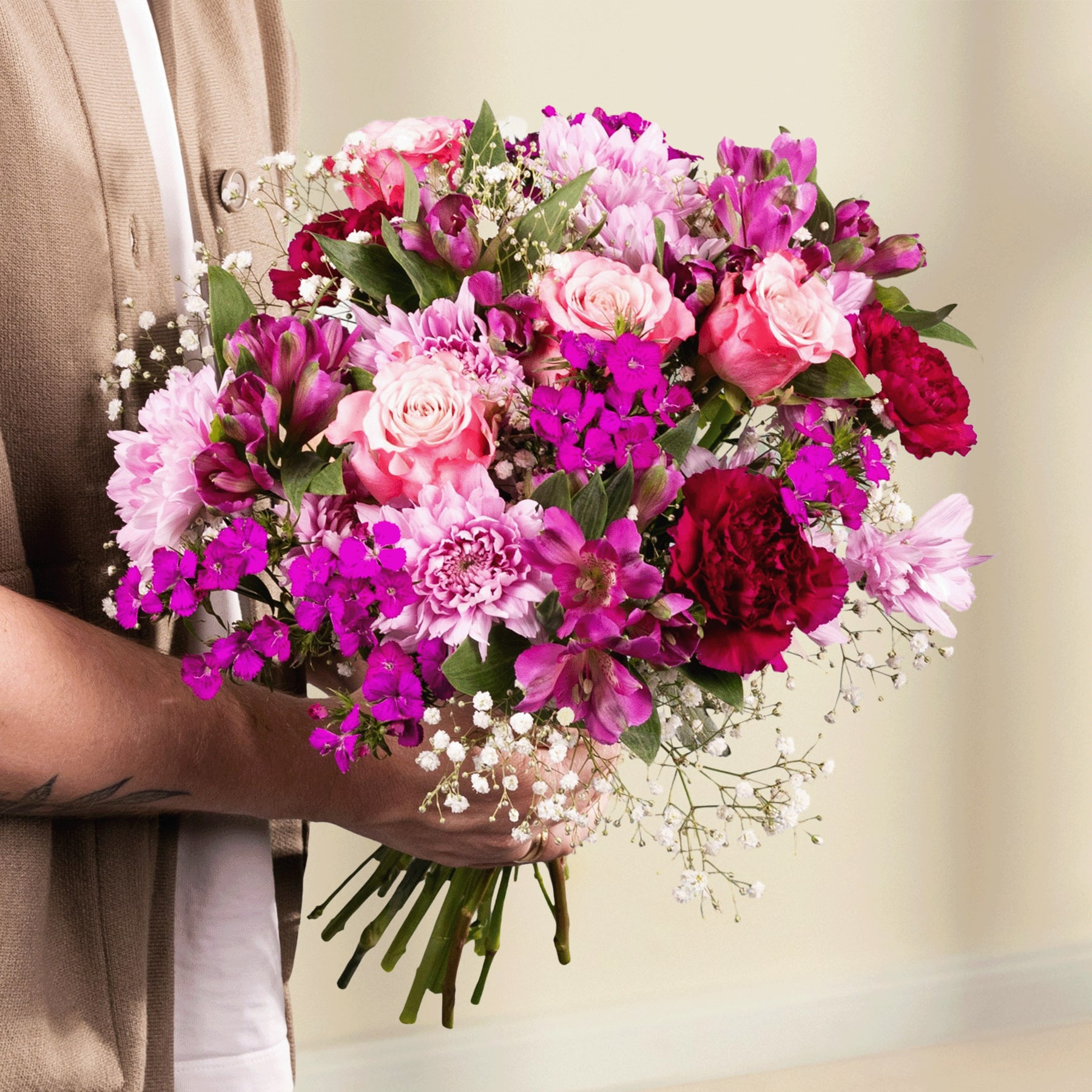 Ein leuchtender Strauß rosa, roter und violetter Blumen, gehalten von einer Person in einem beigen Mantel vor einem hellblauen Hintergrund.