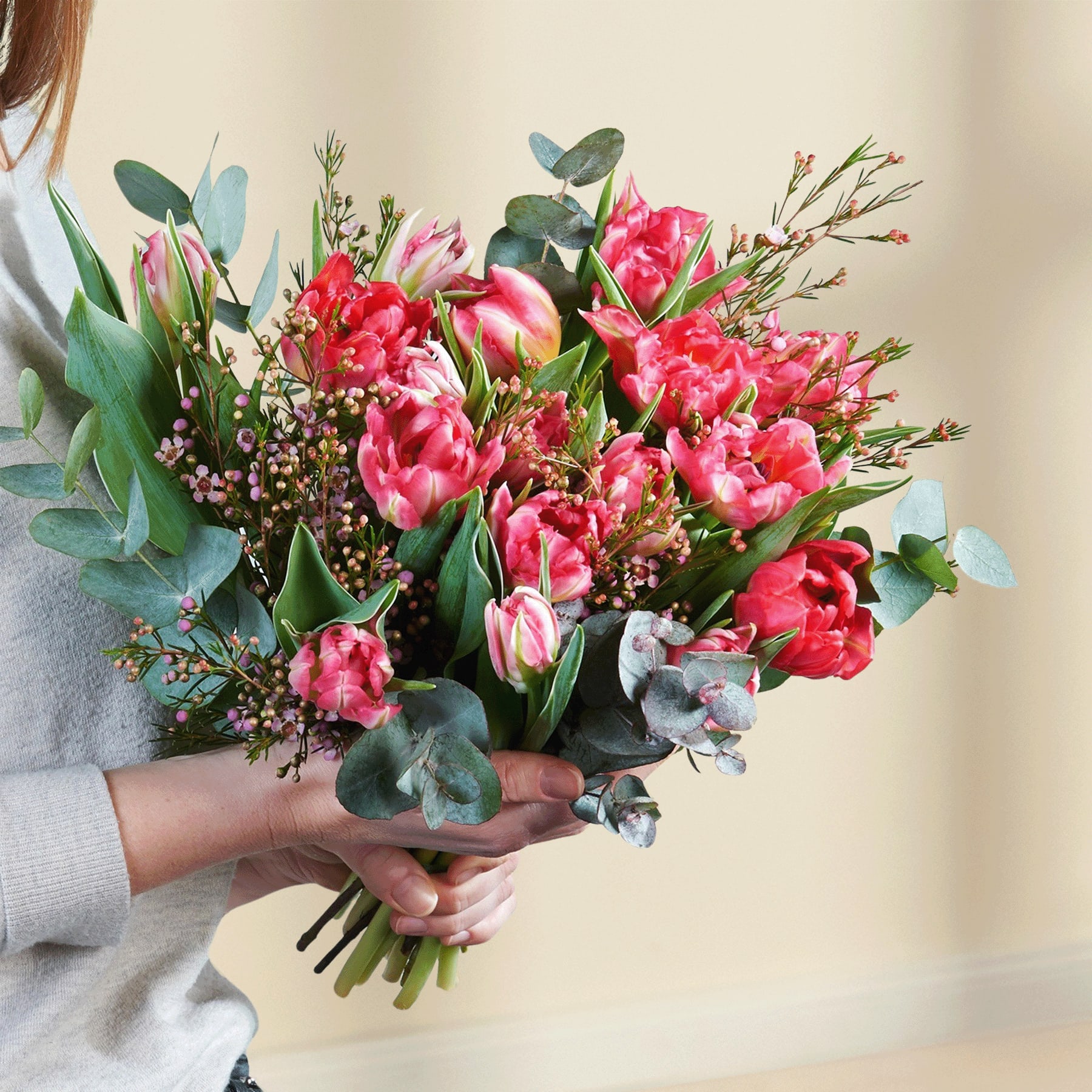 Ein leuchtender Strauß roter und rosa Tulpen, umgeben von üppigem Grün, wird von einer Person in einem grauen Pullover vor einem verschwommenen Hintergrund in den Händen gehalten.