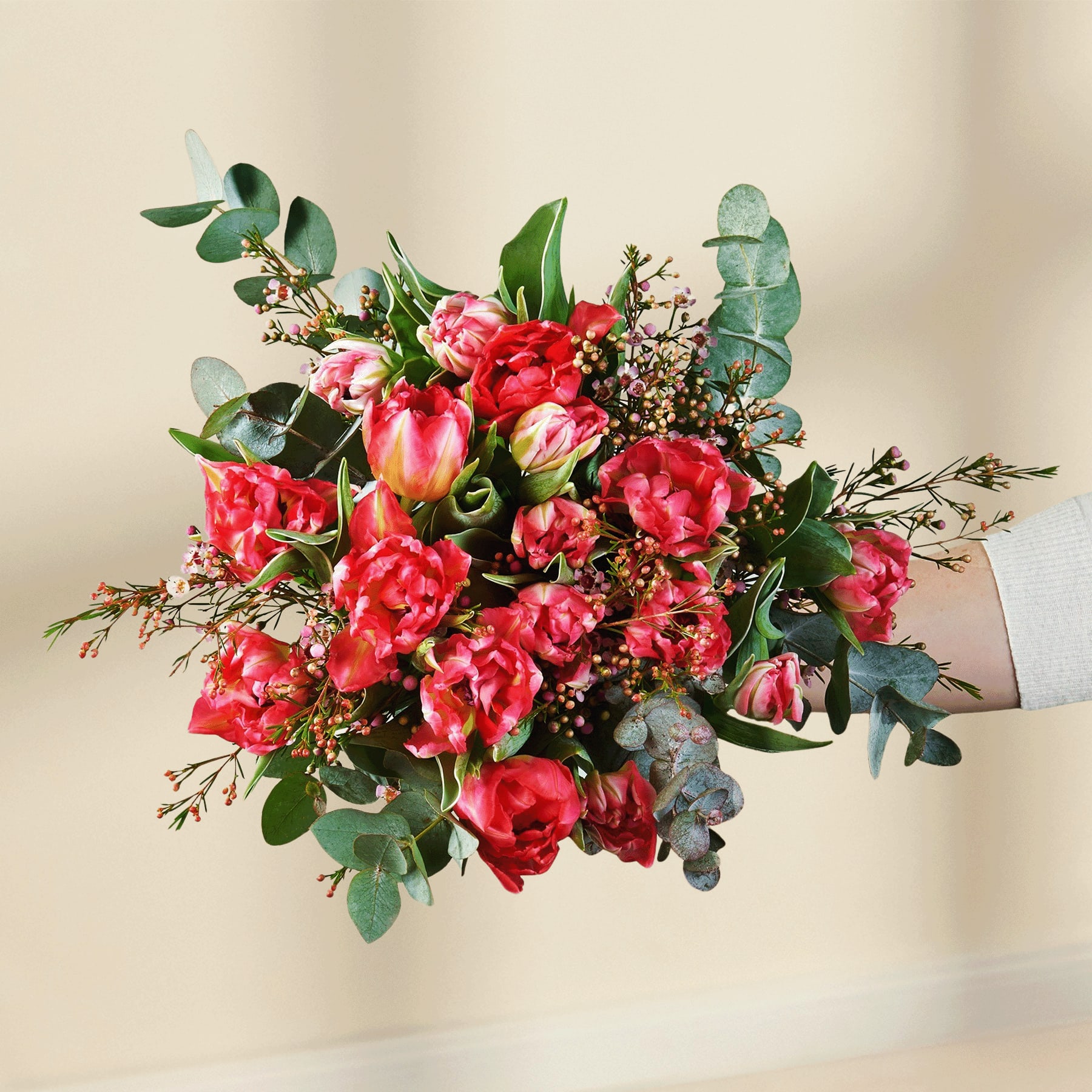Ein leuchtender Strauß üppiger, rosa Pfingstrosen und anderer bunter Blumen, akzentuiert mit Grün, wird vor einem schlichten weißen Hintergrund in den Händen einer Person gehalten.