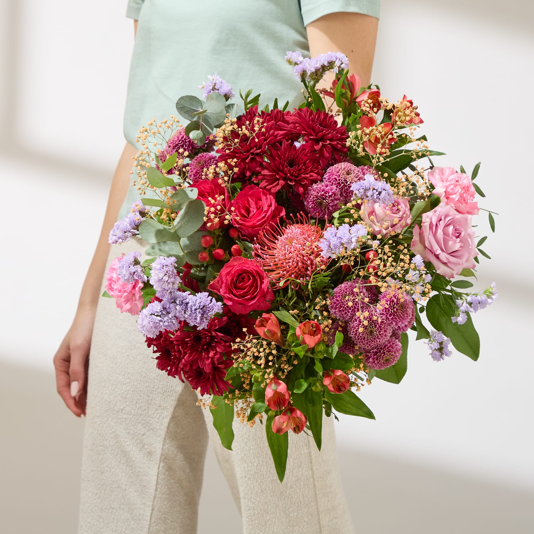 Ein lebhafter und farbenfroher Blumenstrauß aus verschiedenen Blumen, darunter Rosen, Dahlien und Nelken, gehalten von einer Person in einem hellen Hemd vor einem schlichten Hintergrund.