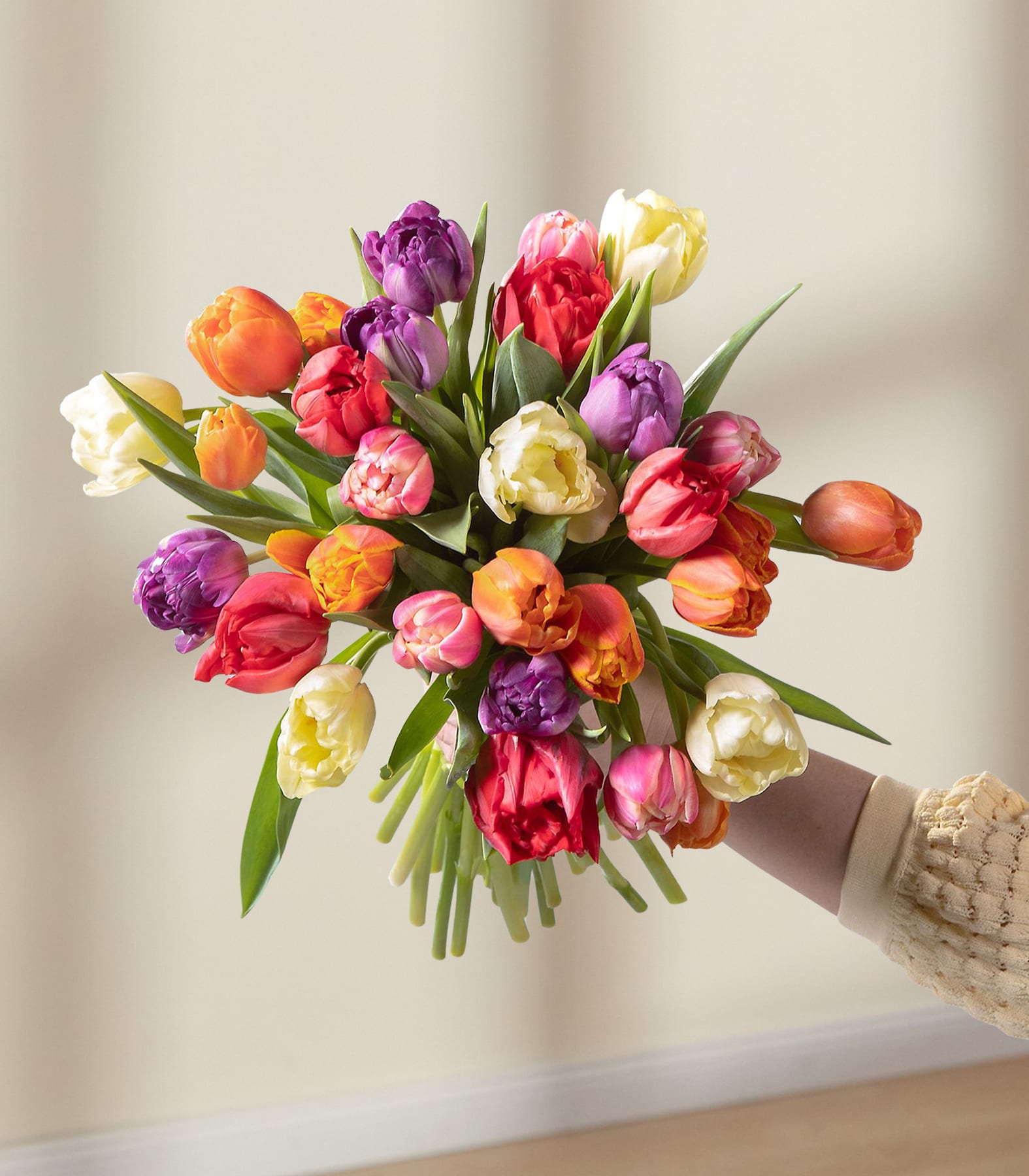 Ein farbenfroher Strauß Tulpen in verschiedenen Rosa-, Rot-, Gelb- und Lilatönen wird von einer Person in den Händen gehalten, vor einem schlichten, hellen Hintergrund.
