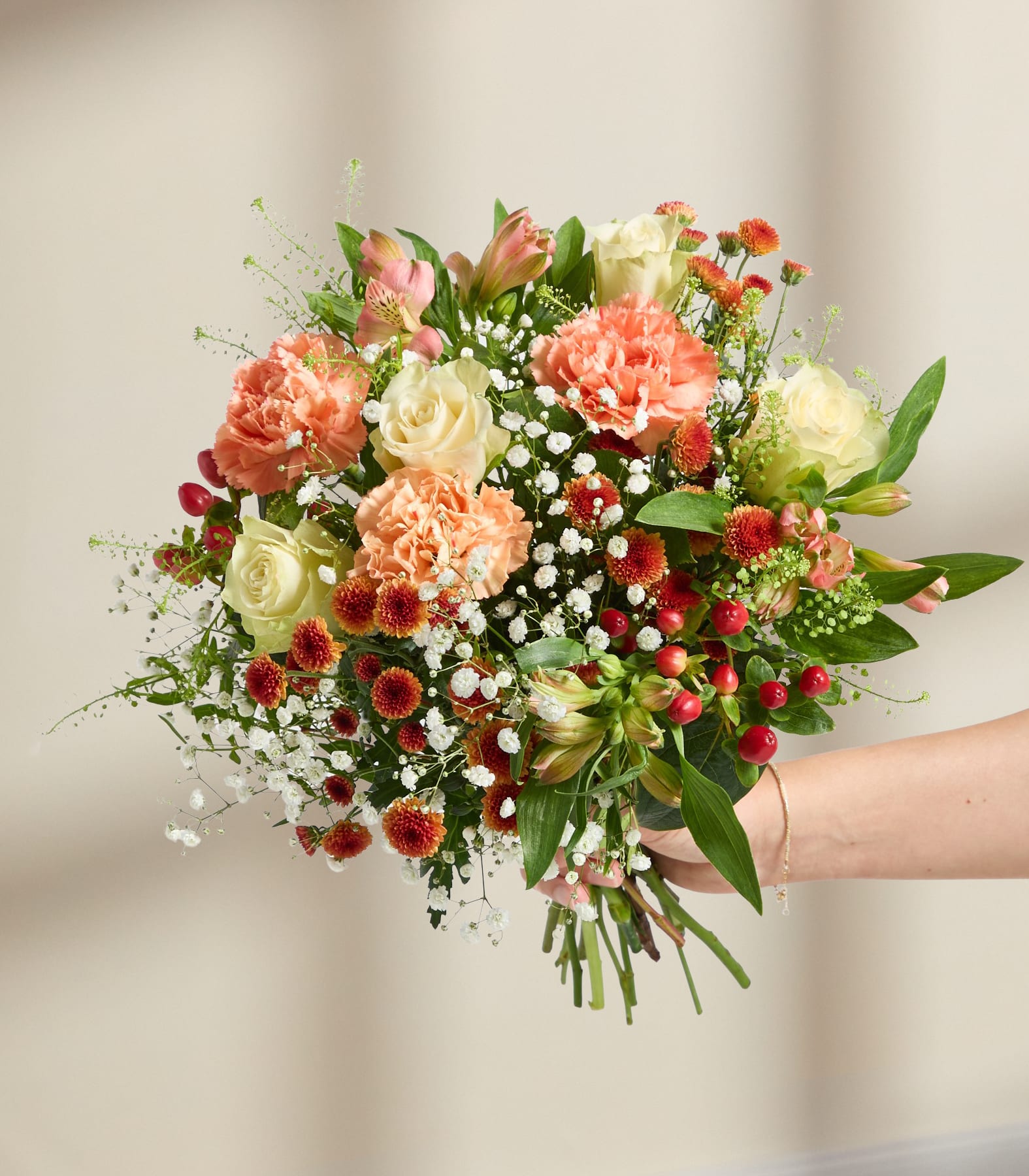 Ein farbenfroher Blumenstrauß aus verschiedenen Blumen, darunter Nelken, Rosen und Grünzeug, der von einer Person in den Händen gehalten wird, vor einem schlichten, hellen Hintergrund.