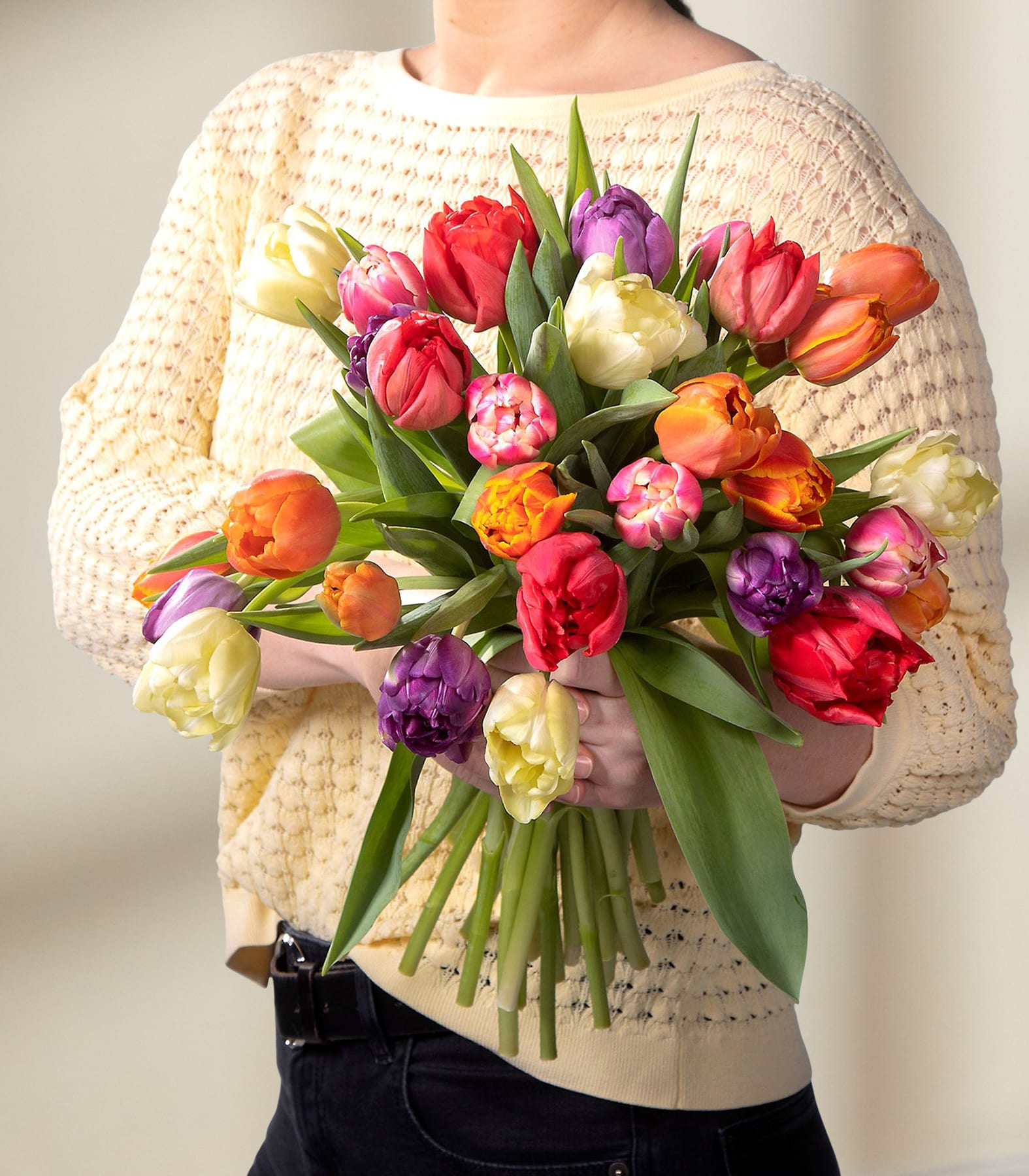 Im Vordergrund wird ein farbenfroher Strauß aus Tulpen und anderen Frühlingsblumen gehalten, im Hintergrund ist eine Person in einem cremefarbenen Pullover zu sehen.