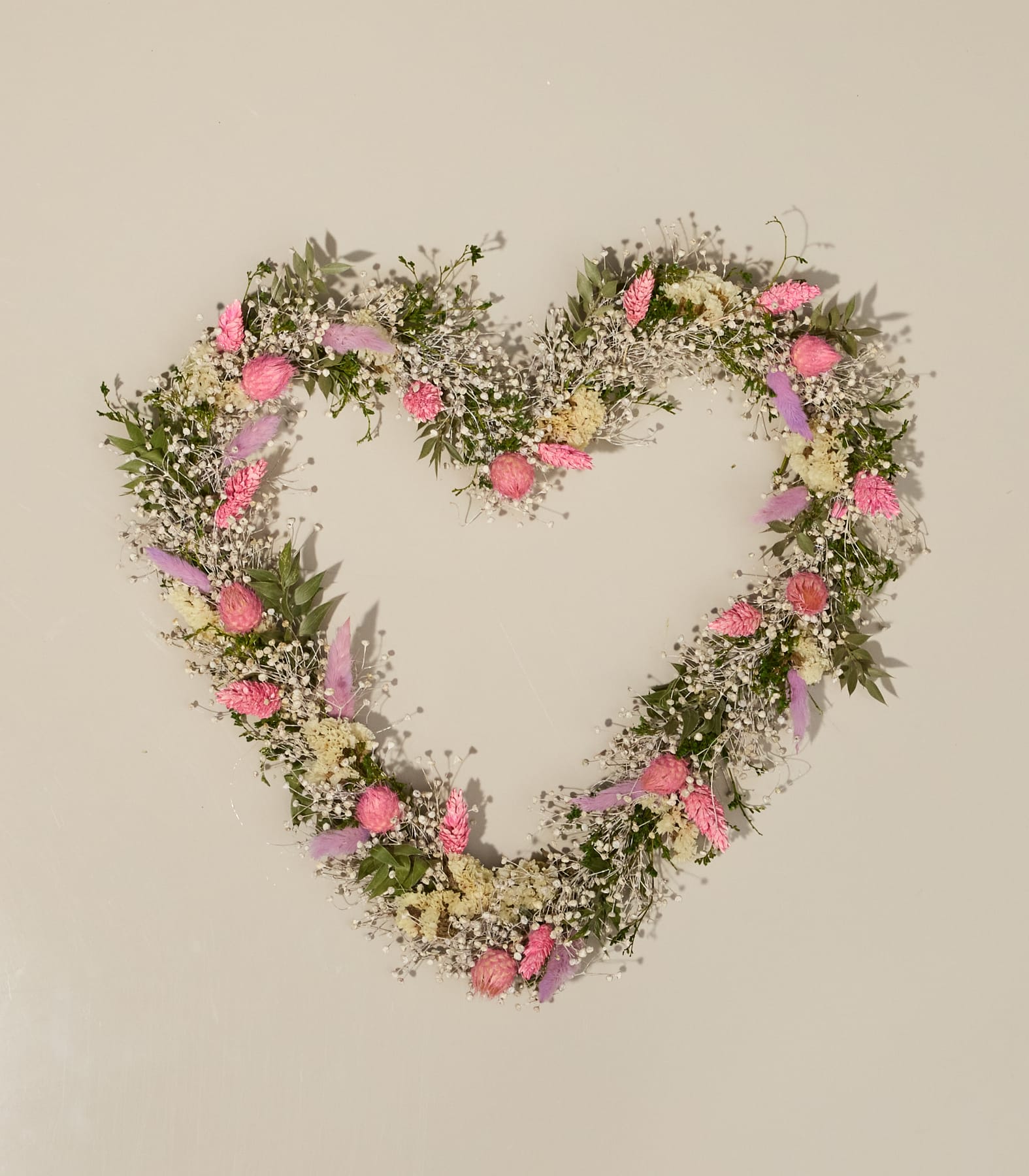 Auf einer schlichten, hellen Fläche liegt ein herzförmiger Kranz aus Trockenblumen und Grünpflanzen.