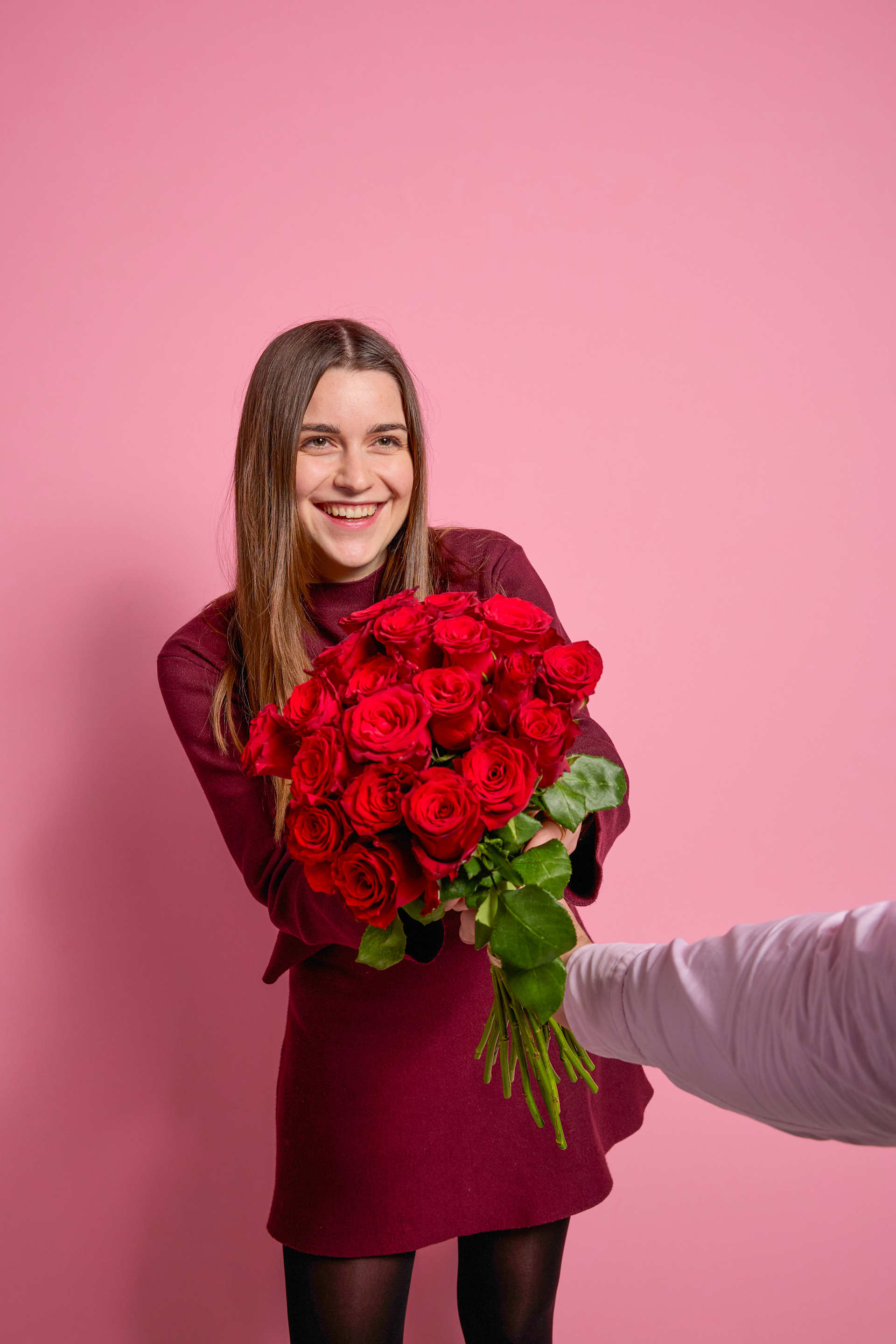 Eine lächelnde junge Frau in einem bordeauxroten Mantel hält einen großen Strauß roter Rosen vor einem leuchtend pinkfarbenen Hintergrund.