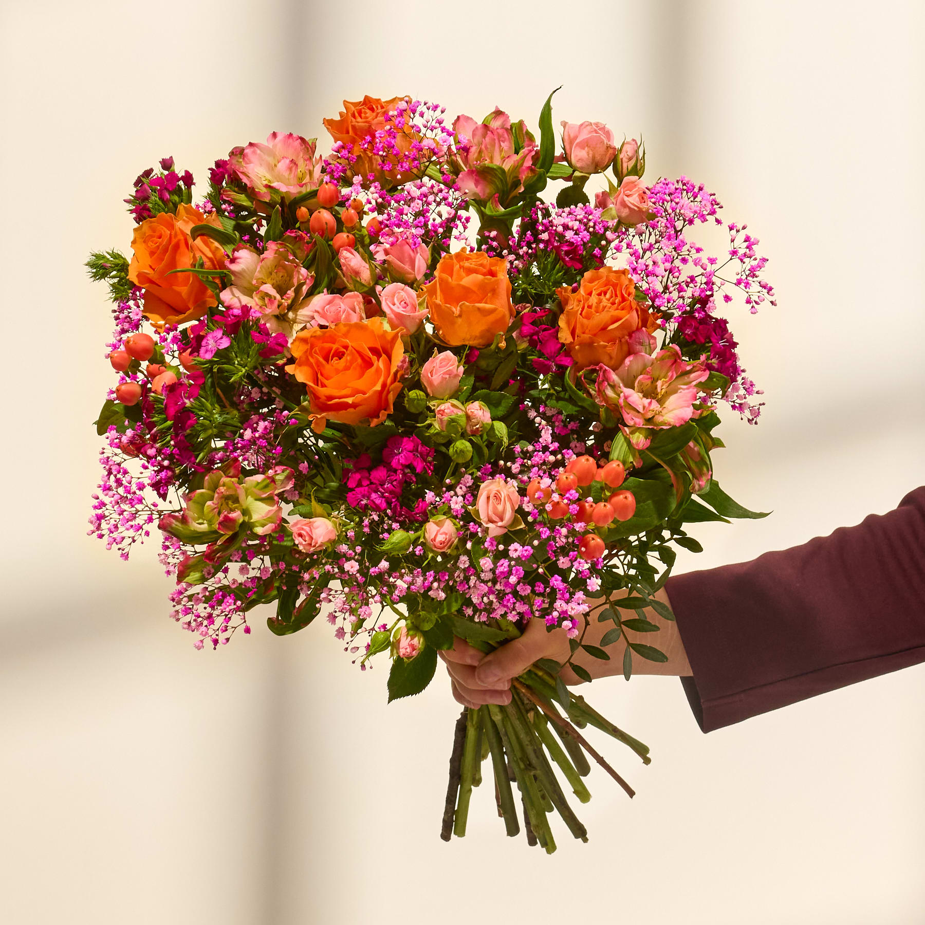 Ein farbenfroher Blumenstrauß, darunter orangefarbene Rosen, rosa Nelken und lila Strandflieder, in den Händen einer Person vor einem schlichten Hintergrund.