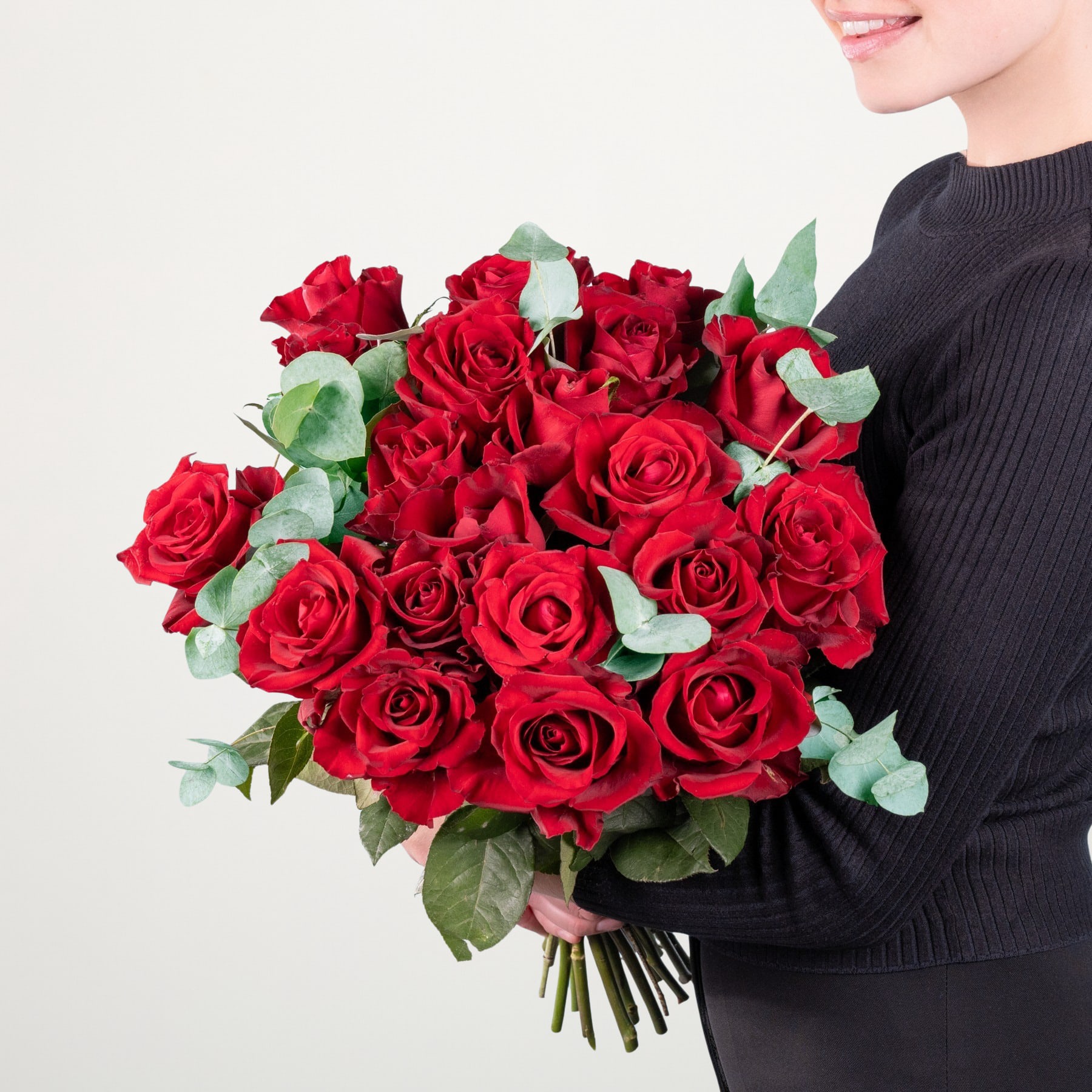 Ein großer Strauß leuchtend roter Rosen, gehalten von einer Person in einem schwarzen Pullover vor einem schlichten weißen Hintergrund.