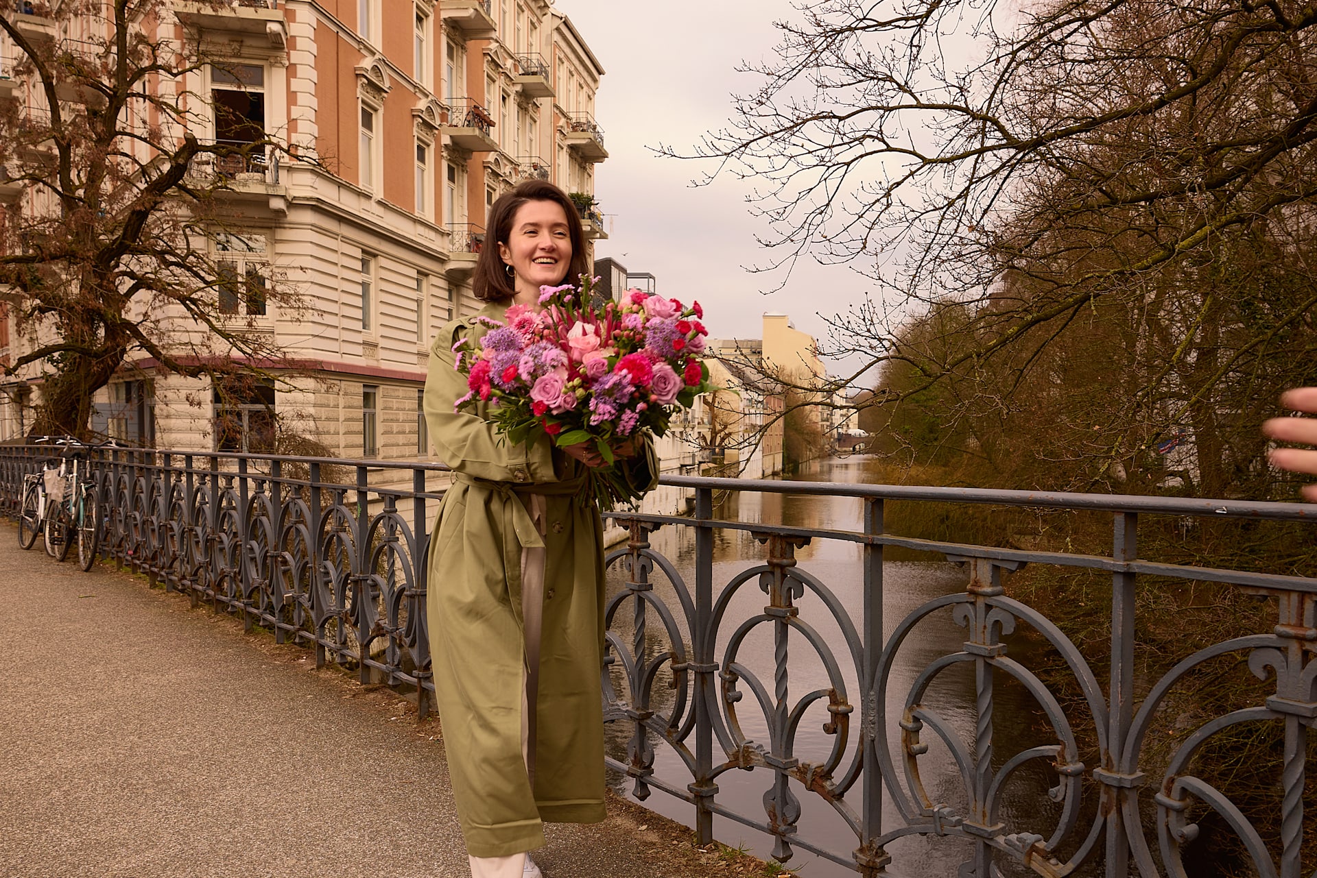 Eine Frau in einem grünen Mantel steht auf einer Brücke und hält einen Strauß leuchtend pinkfarbener Blumen; im Hintergrund sind ein historisches Gebäude und kahle Bäume zu sehen.
