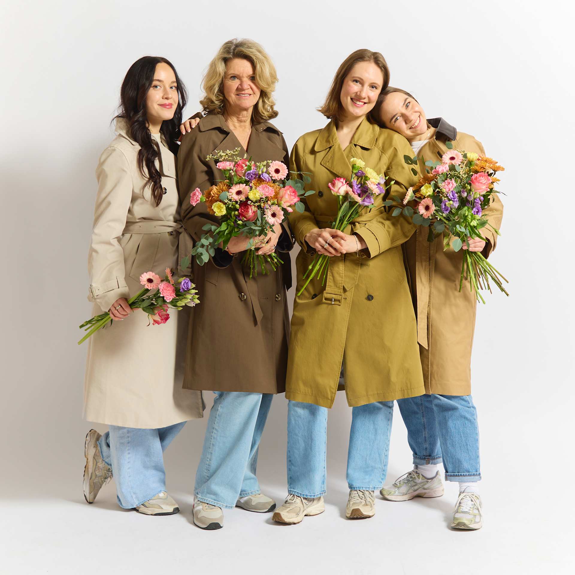 Vier Frauen in legerer Kleidung stehen zusammen, jede hält einen Strauß bunter Blumen vor einem schlichten weißen Hintergrund.