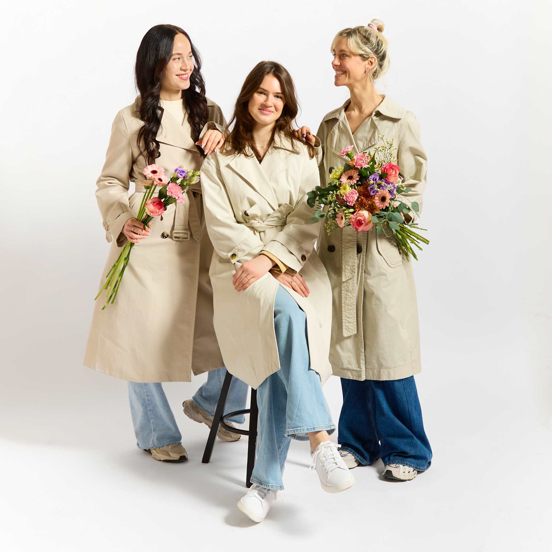 Drei Frauen in lässiger, stilvoller Kleidung stehen zusammen und halten bunte Blumensträuße vor einem schlichten weißen Hintergrund.