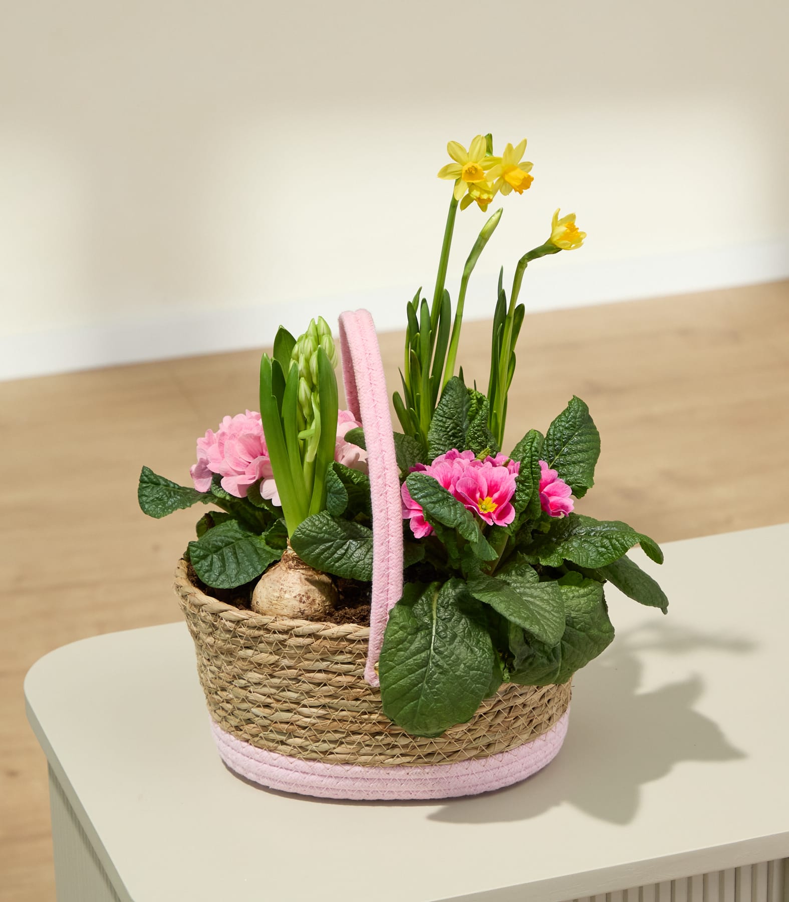 Ein geflochtener Korb gefüllt mit leuchtend violetten und rosa Blumen, darunter eine Hyazinthe, umgeben von grünem Laub, ruht auf einer weißen Oberfläche.