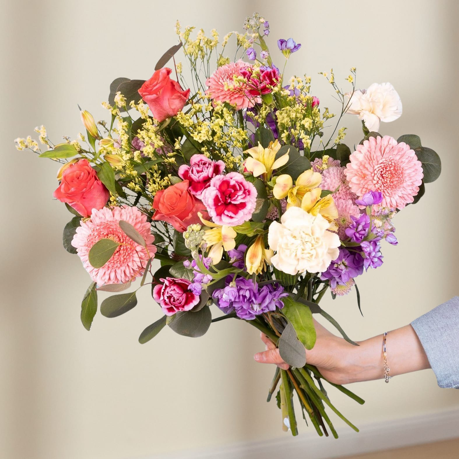 Ein leuchtender Strauß bunter Blumen, darunter Rosen, Nelken und Gänseblümchen, wird vor einem schlichten weißen Hintergrund in den Händen einer Person gehalten.