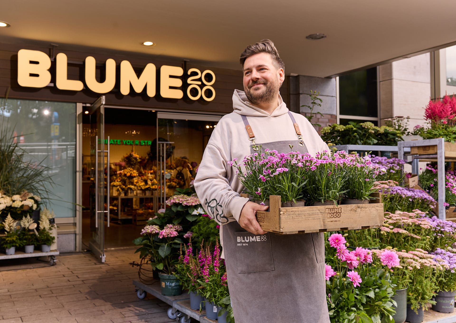 Ein Mann in einer Schürze steht vor einem Blumenladen namens „Blume 2020“ und hält eine Kiste mit bunten Blumen.