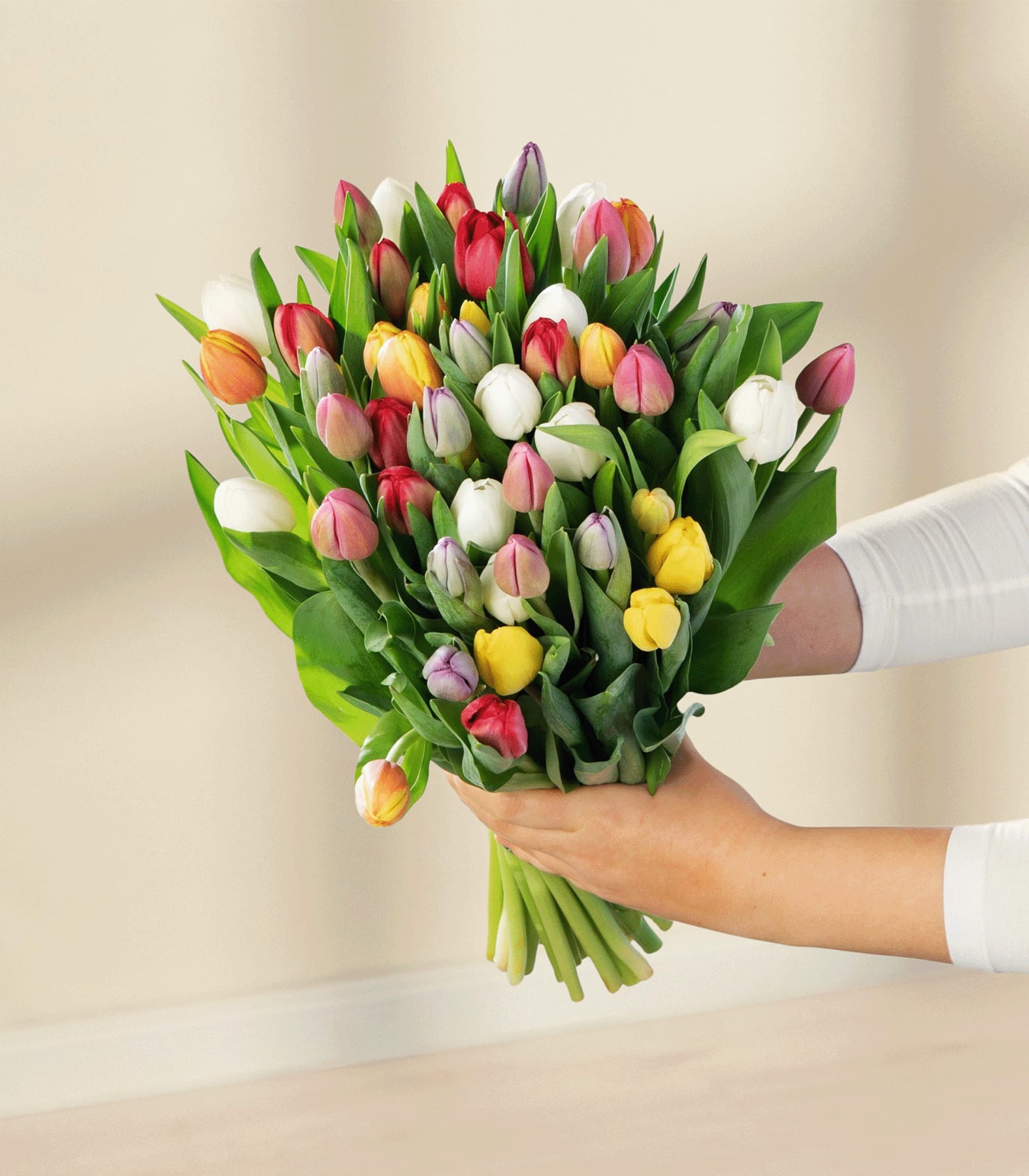 Eine lächelnde Frau in einem weißen Oberteil hält einen leuchtenden Strauß bunter Tulpen vor einem schlichten weißen Hintergrund.