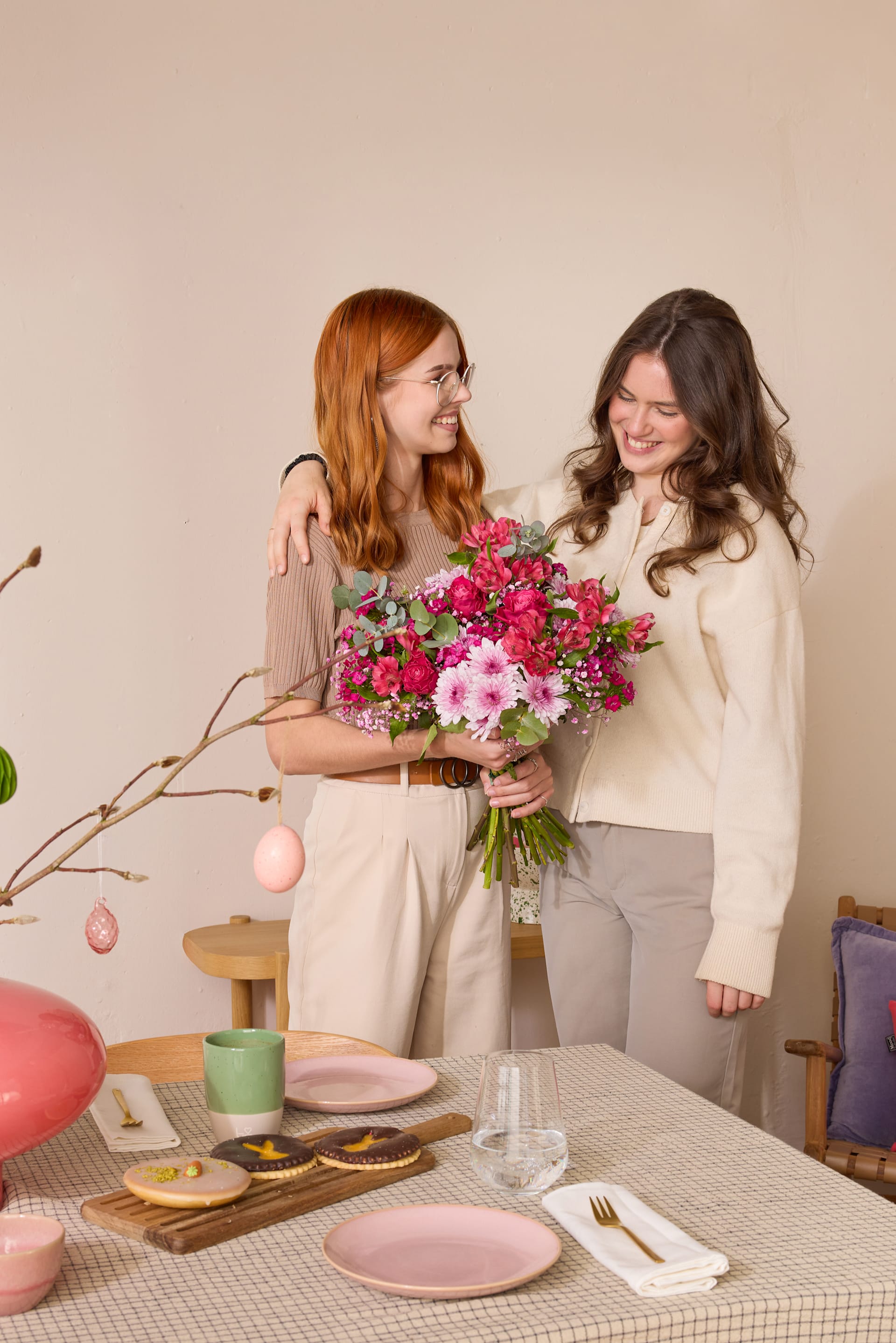 Zwei Frauen in Freizeitkleidung stehen beieinander und halten einen Strauß bunter Blumen. Das Bild ist vor einem schlichten beigen Hintergrund aufgenommen; im Vordergrund ist ein Tisch mit verschiedenen Dekorationsgegenständen zu sehen.