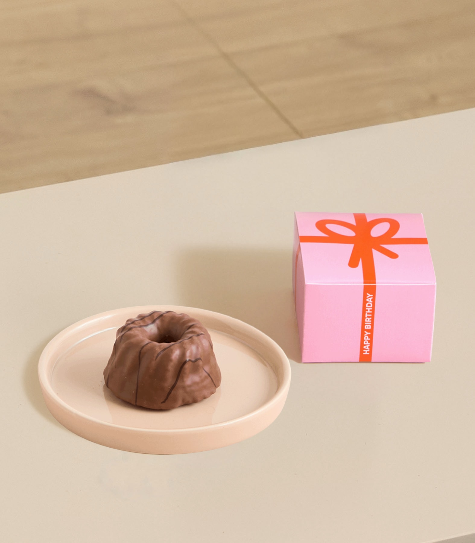 Auf einem blassrosa Teller steht ein kleiner, runder Schokoladenkuchen, im Hintergrund eine grün-weiße Schachtel mit der Aufschrift „Kuchen für dich“.