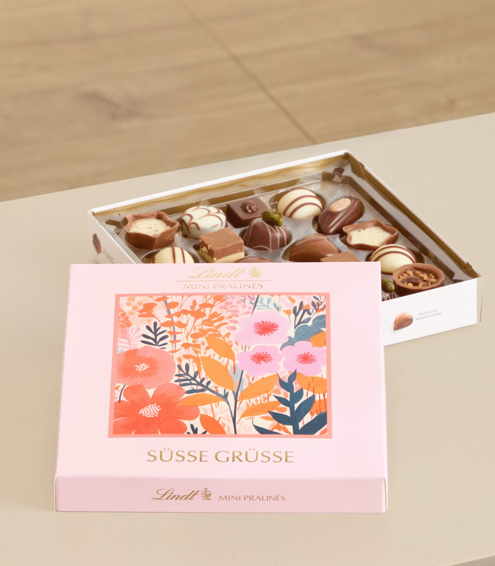 Eine rosa Geschenkbox mit einem Etikett mit Blumenmuster steht auf einem Holztablett, das mit einer Auswahl an Pralinen gefüllt ist.