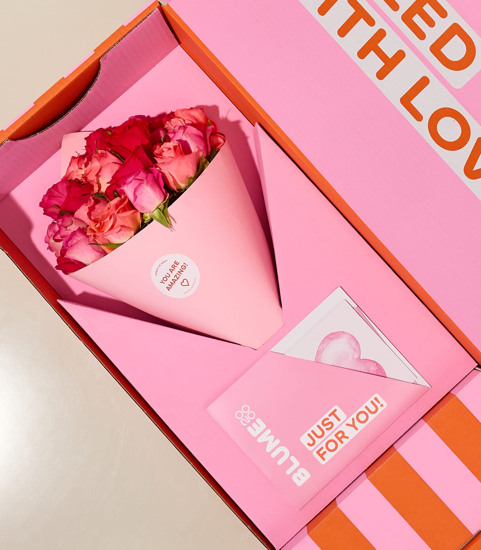 Auf einem Tisch steht eine rosafarbene Schachtel mit der Aufschrift „Voller Liebe“, umgeben von einem Strauß leuchtend roter Rosen in einer Glasvase.