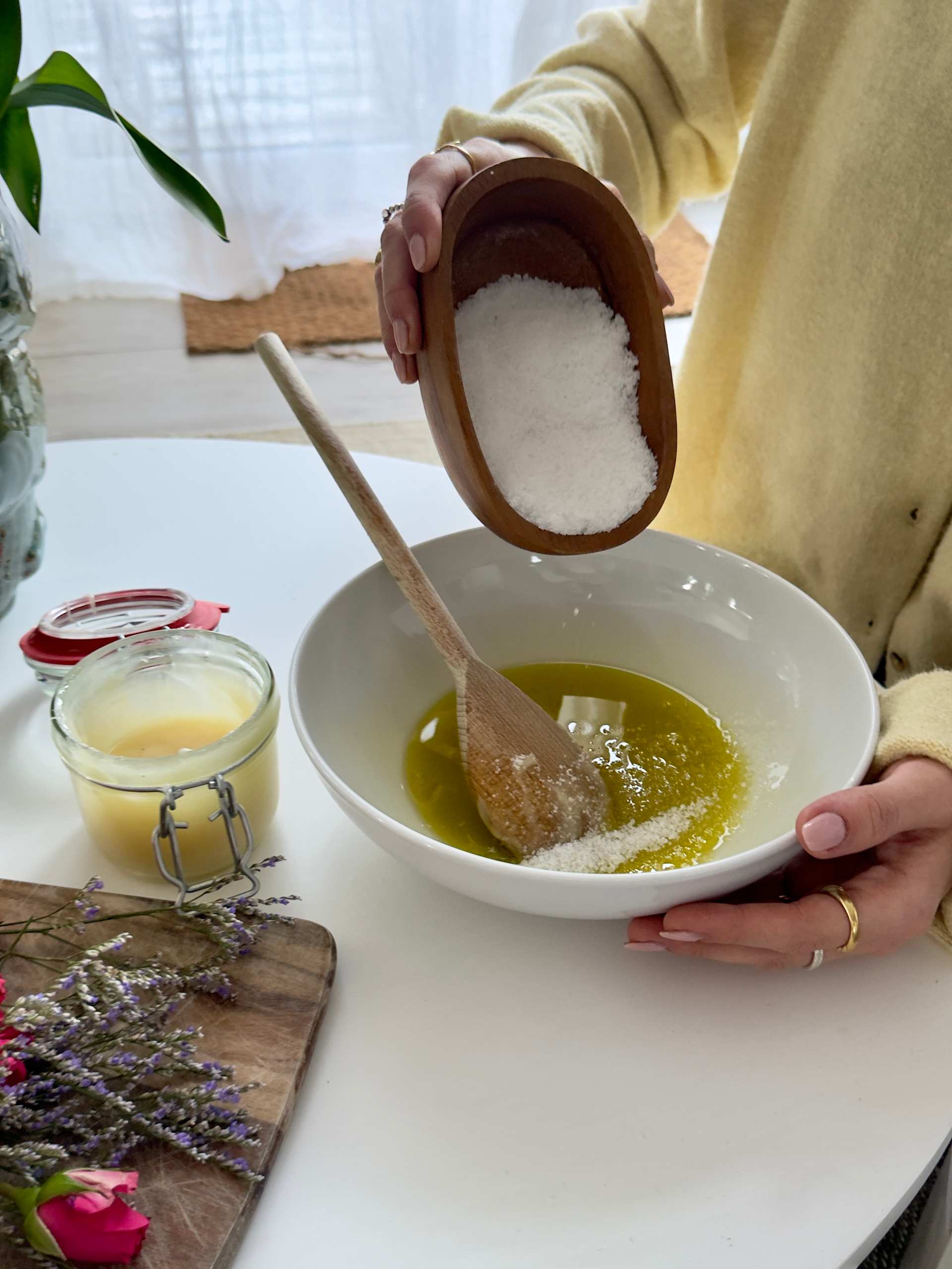Eine Person streut mit den Händen Salz in eine Schüssel mit Flüssigkeit; im Hintergrund sind ein Glas Honig und ein Lavendelzweig zu sehen.
