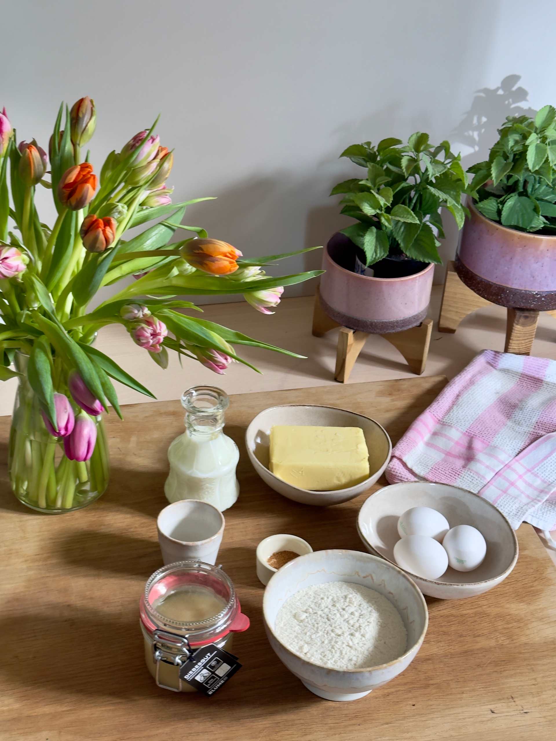 Ein Holztisch mit verschiedenen Küchenutensilien, darunter eine Vase mit Tulpen, Topfpflanzen, eine Schale mit Butter und Schüsseln mit Zutaten wie Reis und Eiern.