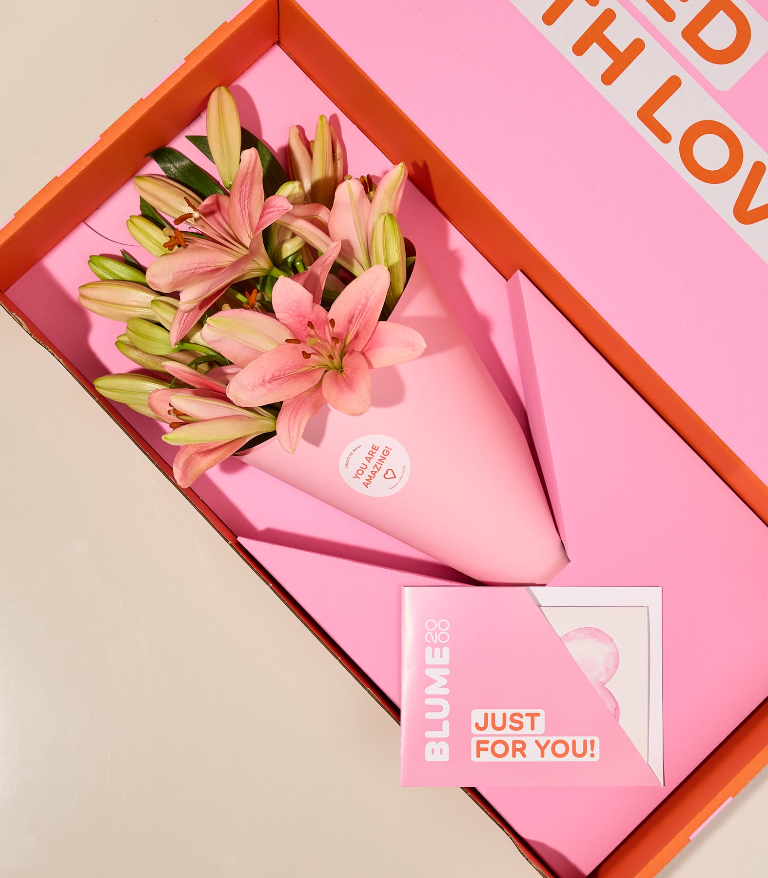 In einer roten Schachtel wird ein Strauß rosa und gelber Lilien präsentiert, dazu eine Grußkarte mit dem Text „Nur für dich!“ vor hellem Hintergrund.