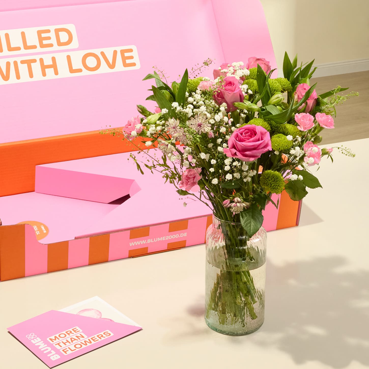Auf einem Tisch steht ein Strauß rosa Rosen und anderer Blumen in einer Glasvase; im Hintergrund ist eine rosa-orangefarbene Geschenkbox mit der Aufschrift „Mit Liebe gefüllt“ zu sehen.