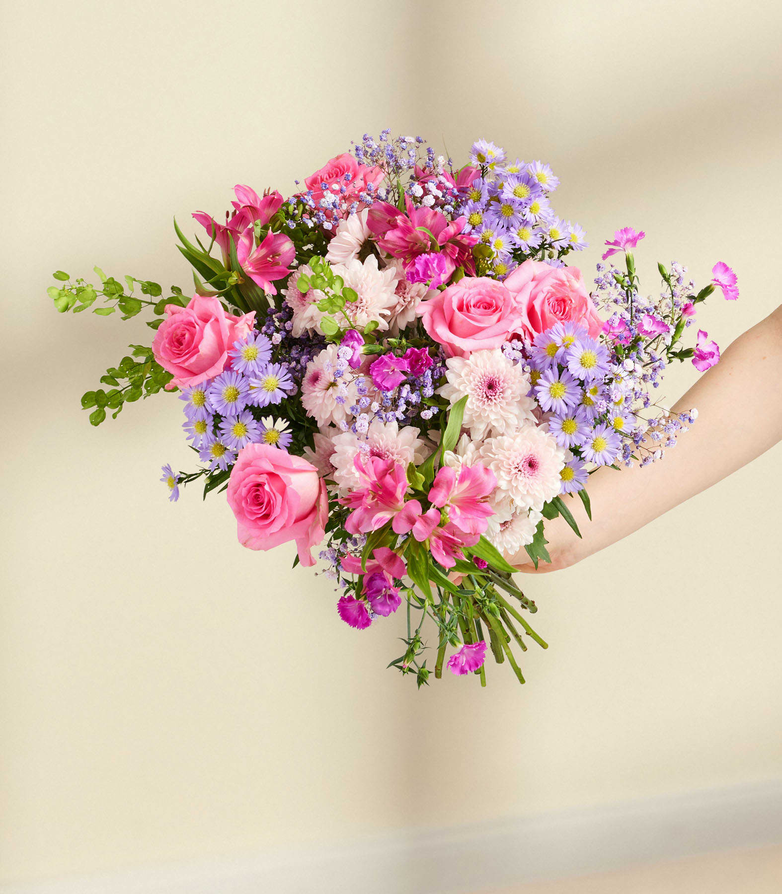 Ein leuchtender Strauß aus rosa Rosen, violetten Blumen und Grünpflanzen steht in einer Glasvase vor einem schlichten weißen Hintergrund.