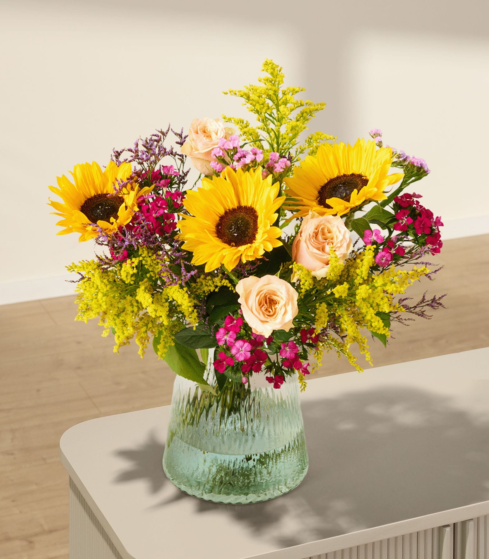 Ein farbenfroher Strauß aus Sonnenblumen, Rosen und anderen bunten Blumen, arrangiert in einer grünen Glasvase, steht auf einer Holzoberfläche vor einem neutralen Hintergrund.
