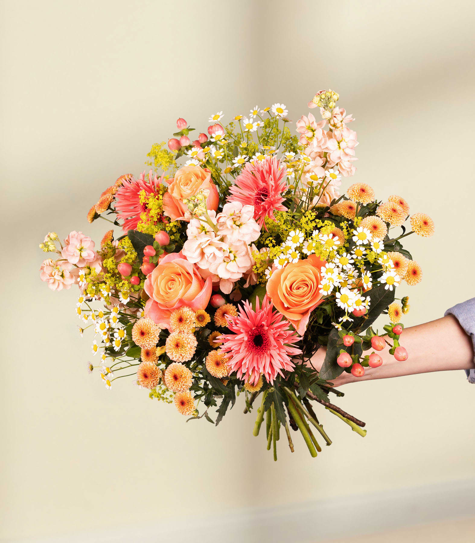 Ein lebhafter Strauß bunter Blumen, darunter rosa und gelbe Rosen, orangefarbene Gerbera und weiße und gelbe Chrysanthemen, in den Händen einer Person vor einem schlichten weißen Hintergrund.