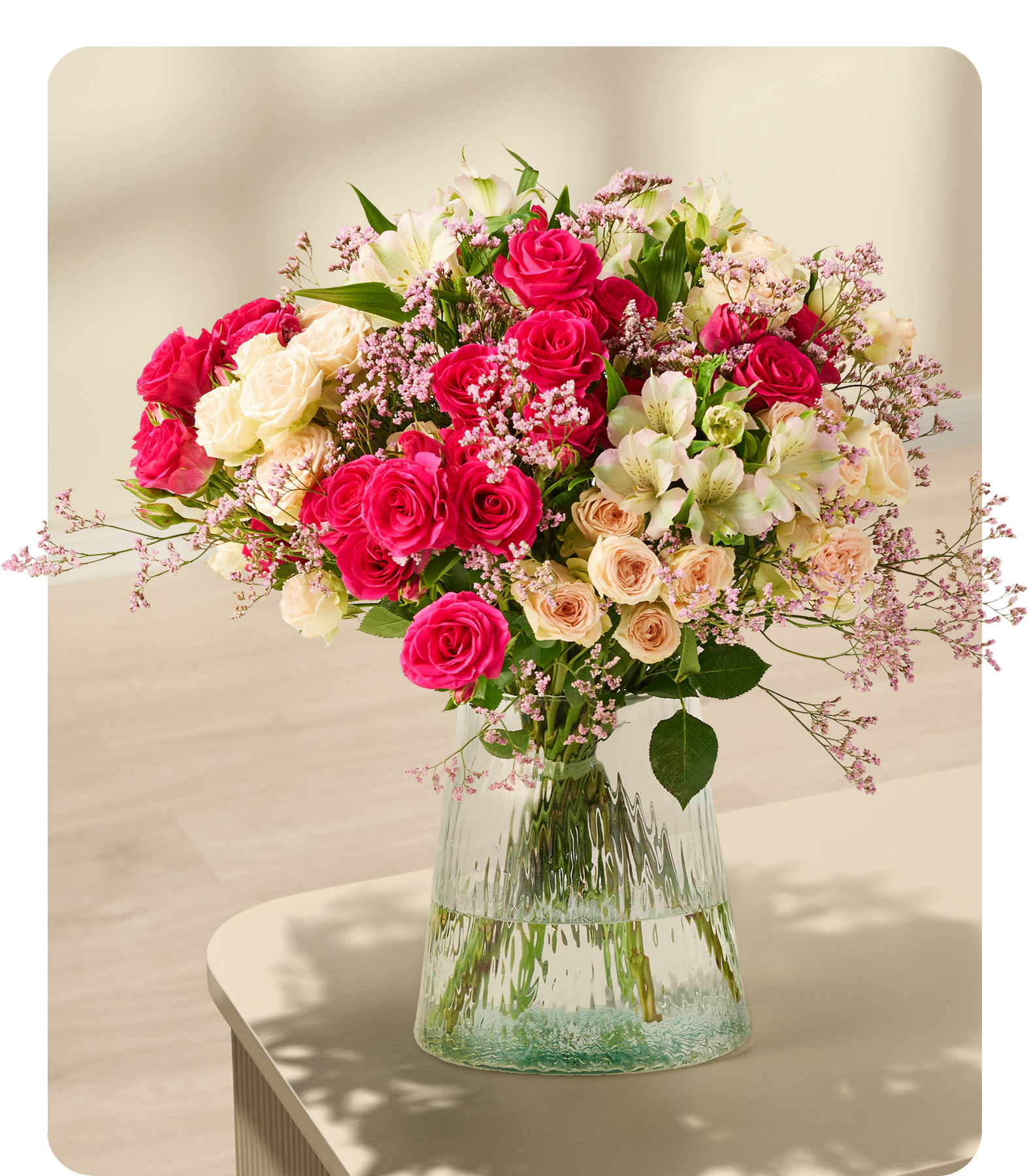 Ein leuchtender Strauß roter Rosen, weißer Nelken und zartrosa Blumen in einer Glasvase steht auf einem mintfarbenen Sockel vor einem schlichten weißen Hintergrund.