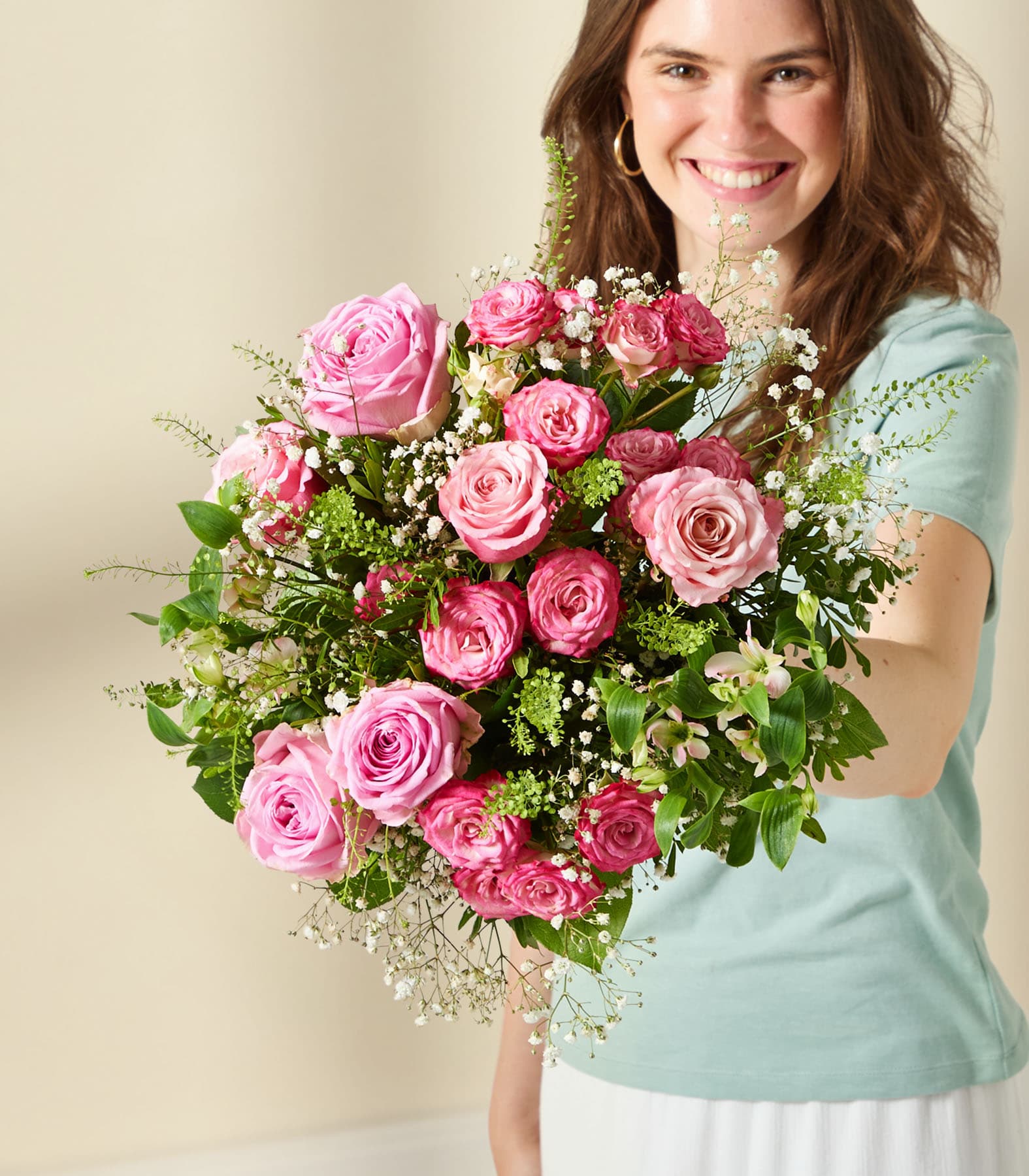 Eine junge Frau mit einem warmen Lächeln hält vor einem neutralen Hintergrund einen leuchtenden Strauß aus rosa Rosen und anderen Blumen.