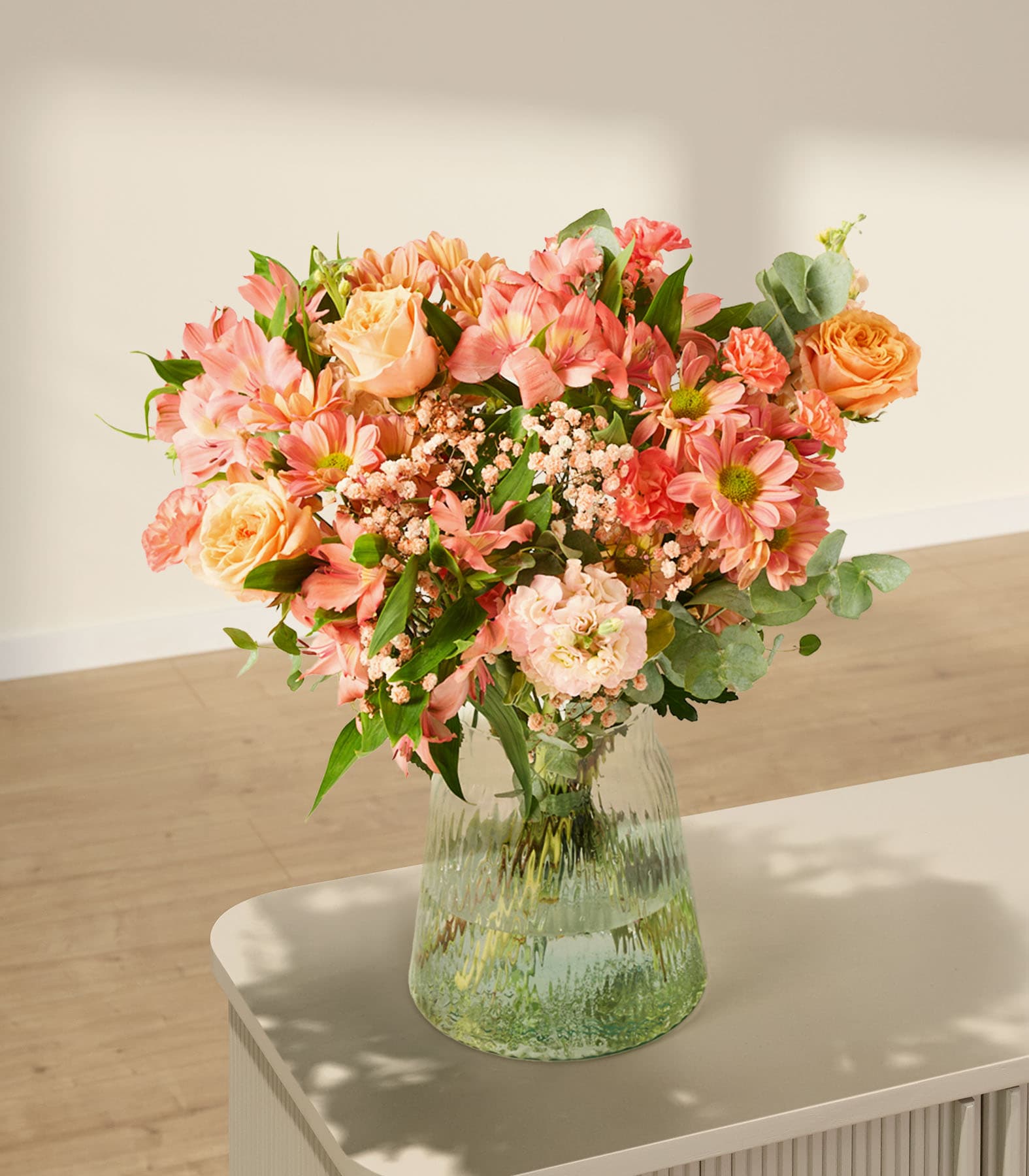 Ein leuchtender Strauß rosa, orangefarbener und gelber Blumen, arrangiert in einer Glasvase vor einem schlichten weißen Hintergrund.